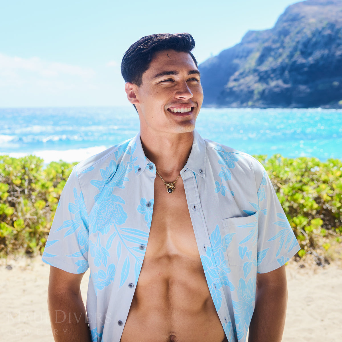 Dane Little wearing a light blue and white patterned Kahala shirt and an Octopus Tahitian black pearl pendant outdoors with ocean and mountains in the background.
