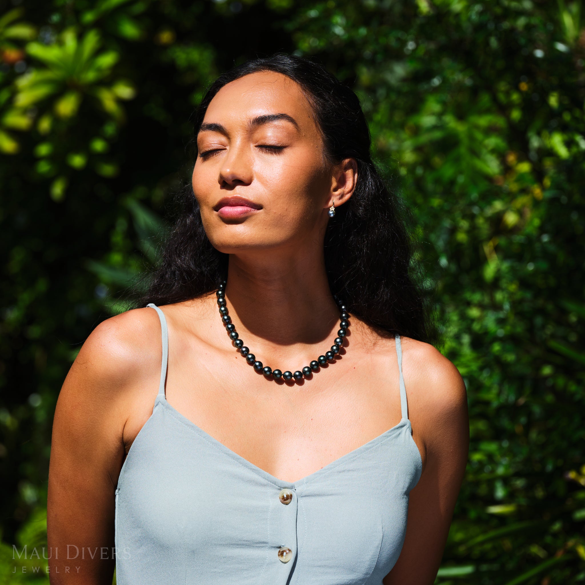 Woman wearing a Tahitian black pearl necklace and plumeria Tahitian black pearl earrings with a green leafy background.