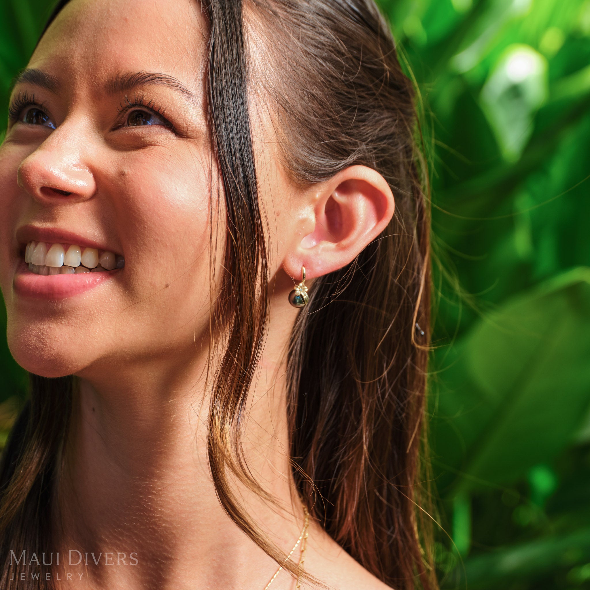 Close-up of a smiling woman wearing a Plumeria Tahitian Black Pearl Earring in 14k yellow gold with diamond accent on her left ear, against a blurred leafy background