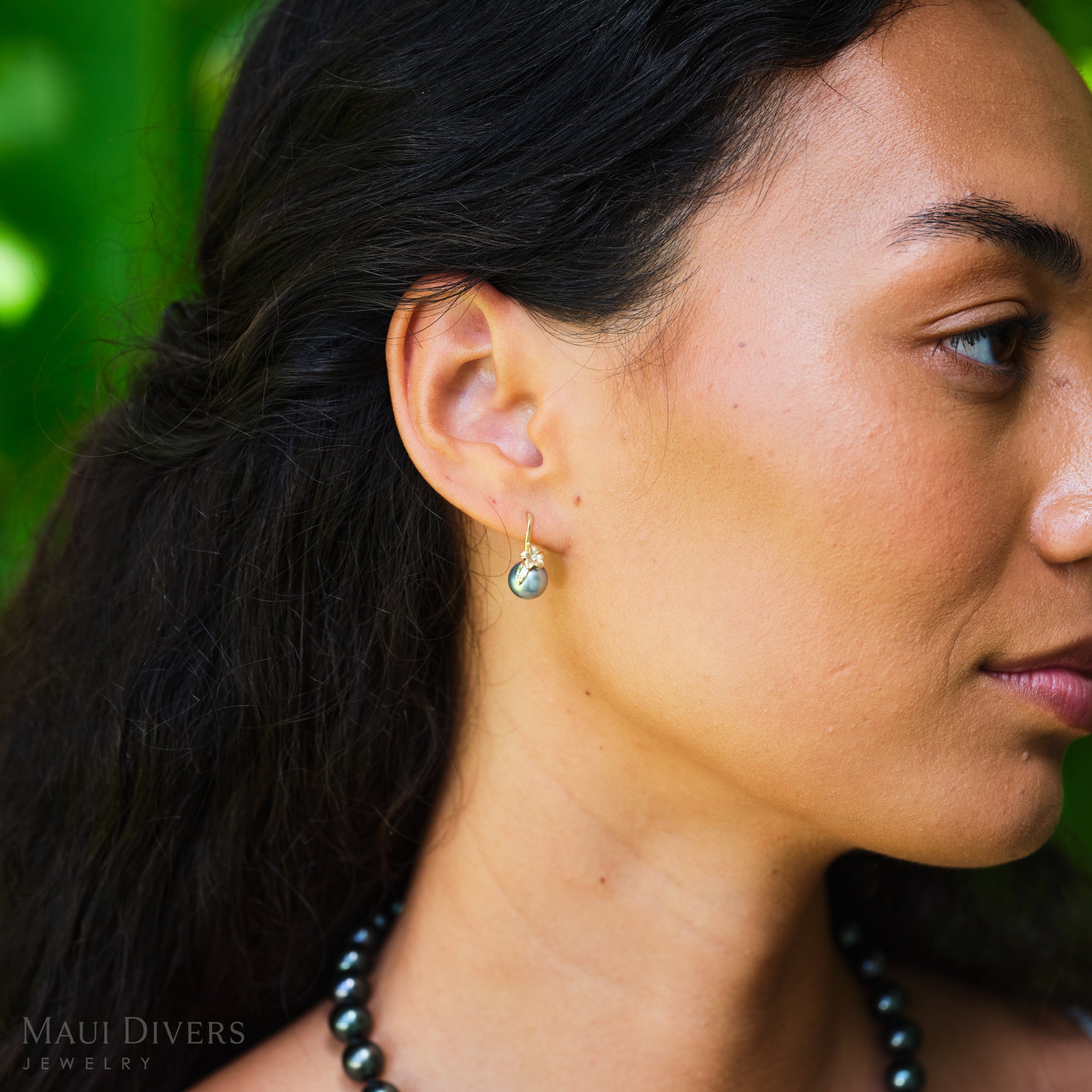 Close-up of a woman wearing plumeria Tahitian black pearl earrings with a blurred green background.