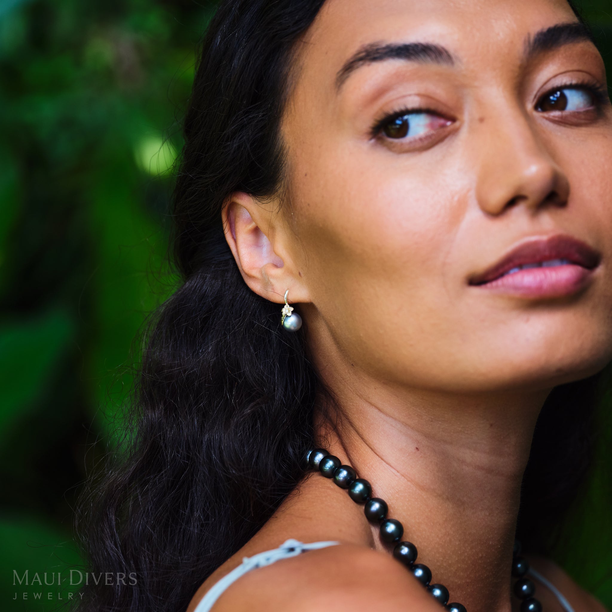 Close-up of a woman wearing a gold plumeria pearl earring with a blurred green background.
