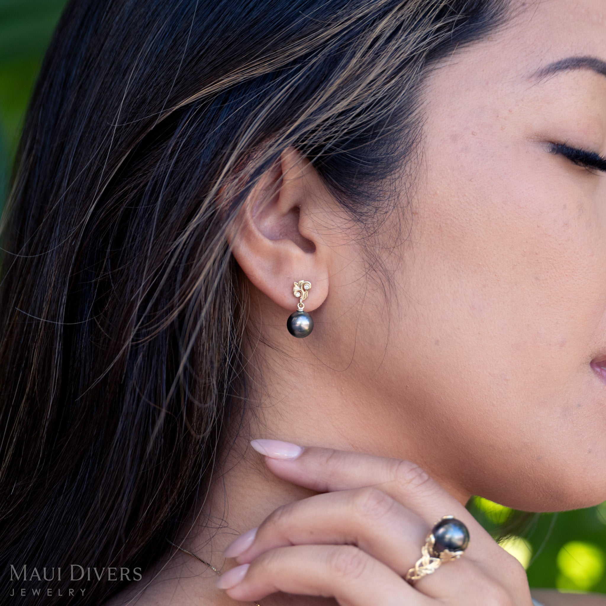 Close-up of a woman wearing black pearl earrings and a matching ring with a blurred green background.