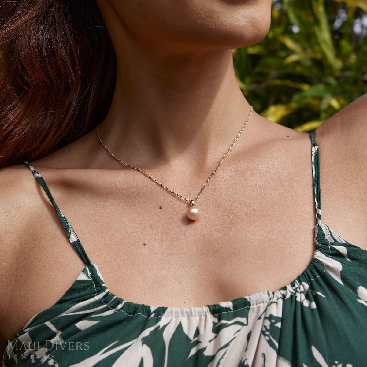 Close-up of a woman in a green top wearing a simple peach/pink pearl pendant with a 14k yellow gold bail on a singapore chain with plants in the background