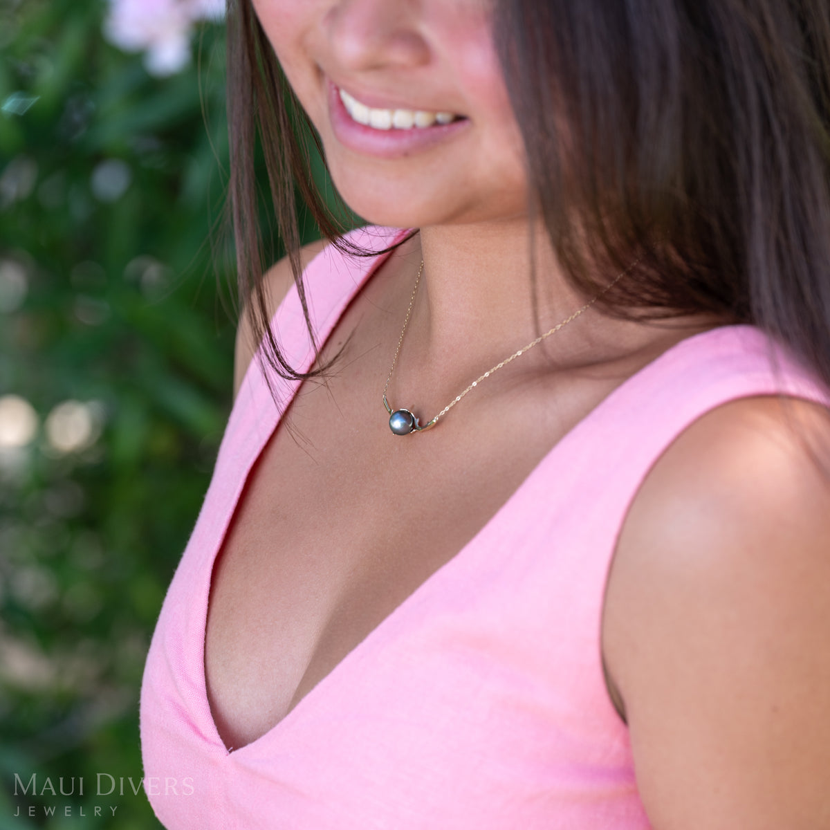 A close-up of a smiling woman in a light pink top wearing a Heritage Tahitian Black Pearl Necklace in 14k yellow gold against a blurred leafy background