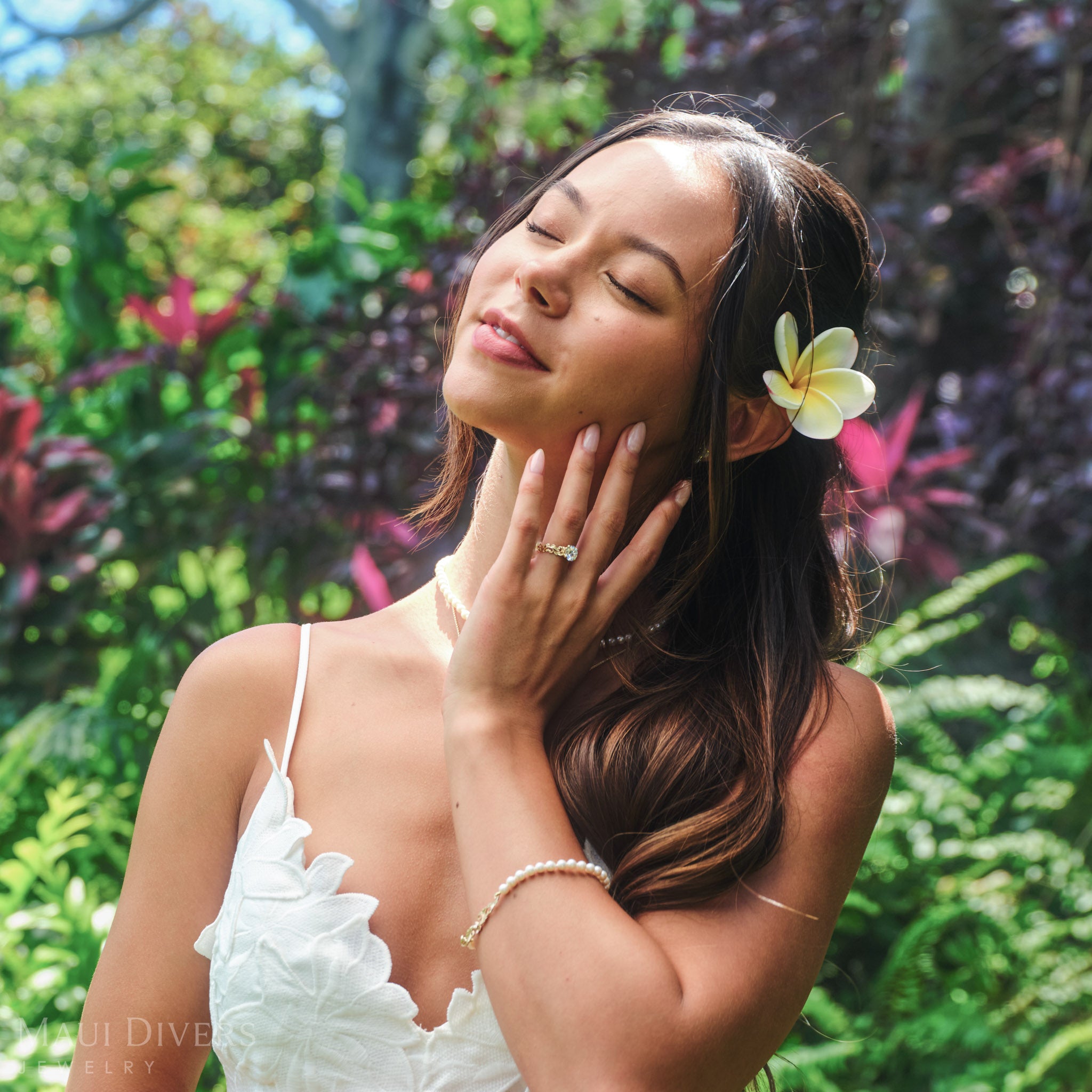 Smiling woman in a white top with a plumeria tucked behind her left ear wearing a Living Heirloom Solitaire Lab Grown Diamond Engagement Ring in 14k yellow gold on her ring finger, against a blurred tropical background