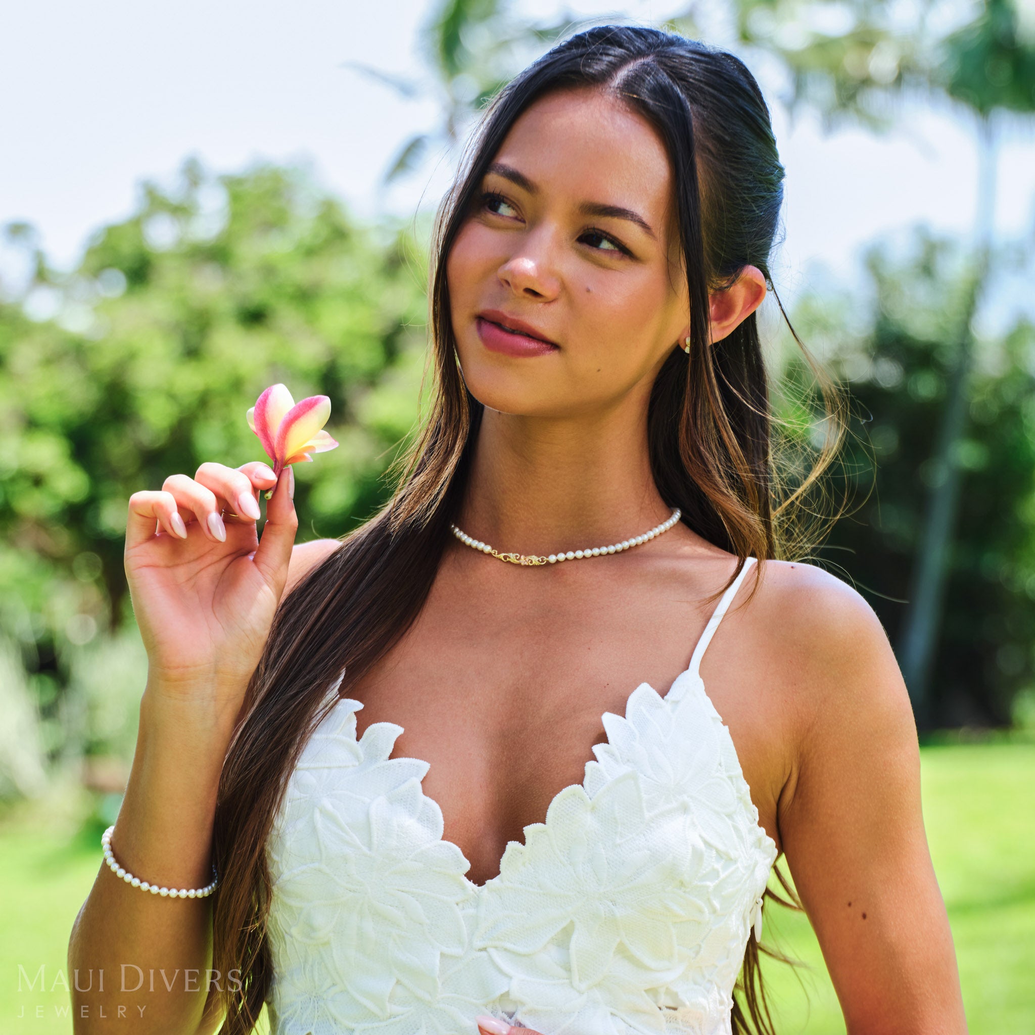 Smiling woman holding a plumeria flower wearing a Hawaiian Heirloom Plumeria Freshwater White Pearl Necklace in Two Tone Gold with Diamond against a blurred park background