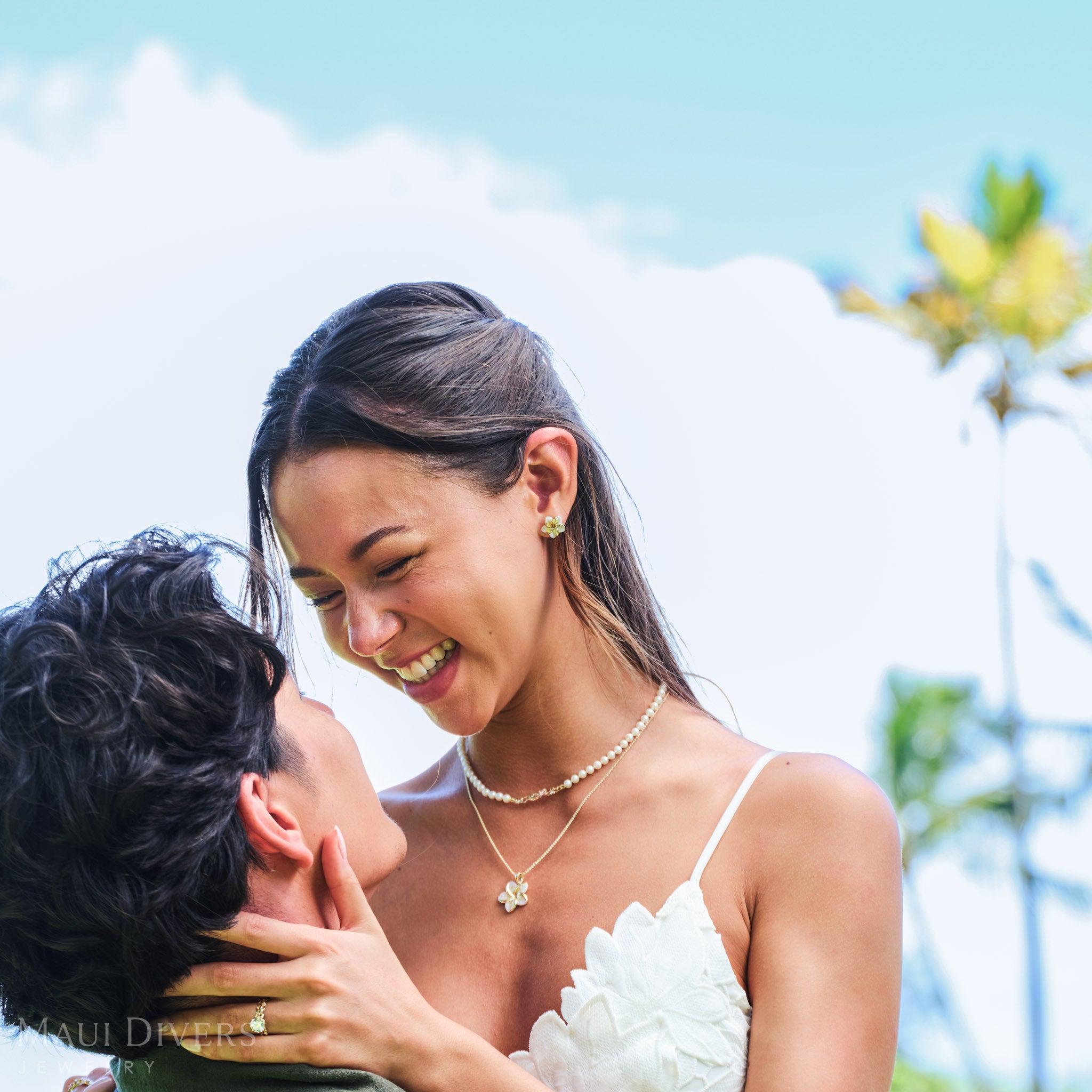 Smiling woman with a plumeria flower behind her ear wearing Plumeria Mother of Pearl earring and pendant in 14k yellow gold and a Hawaiian Heirloom freshwater pearl strand necklace, being lifted by a man against a blurred tropical background