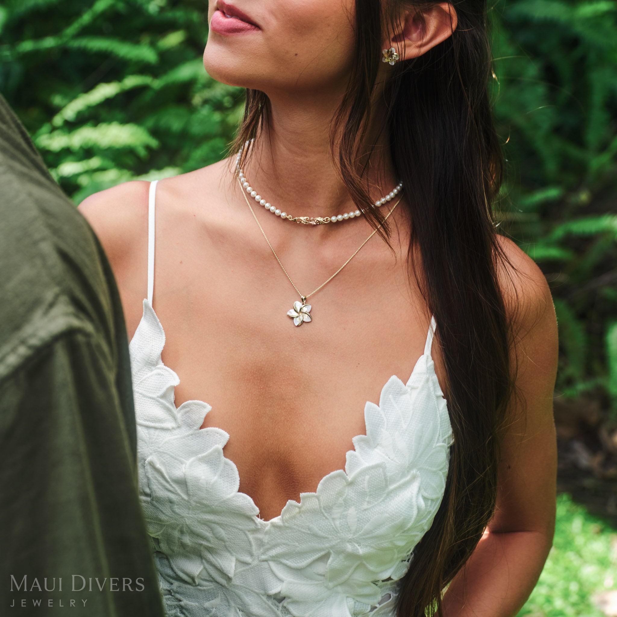 Close-up of a smiling woman in a white dress wearing a Plumeria Mother of Pearl Pendant in 14k yellow gold around her neck, paired with a Hawaiian Heirloom freshwater pearl strand necklace, against a blurred leafy green background