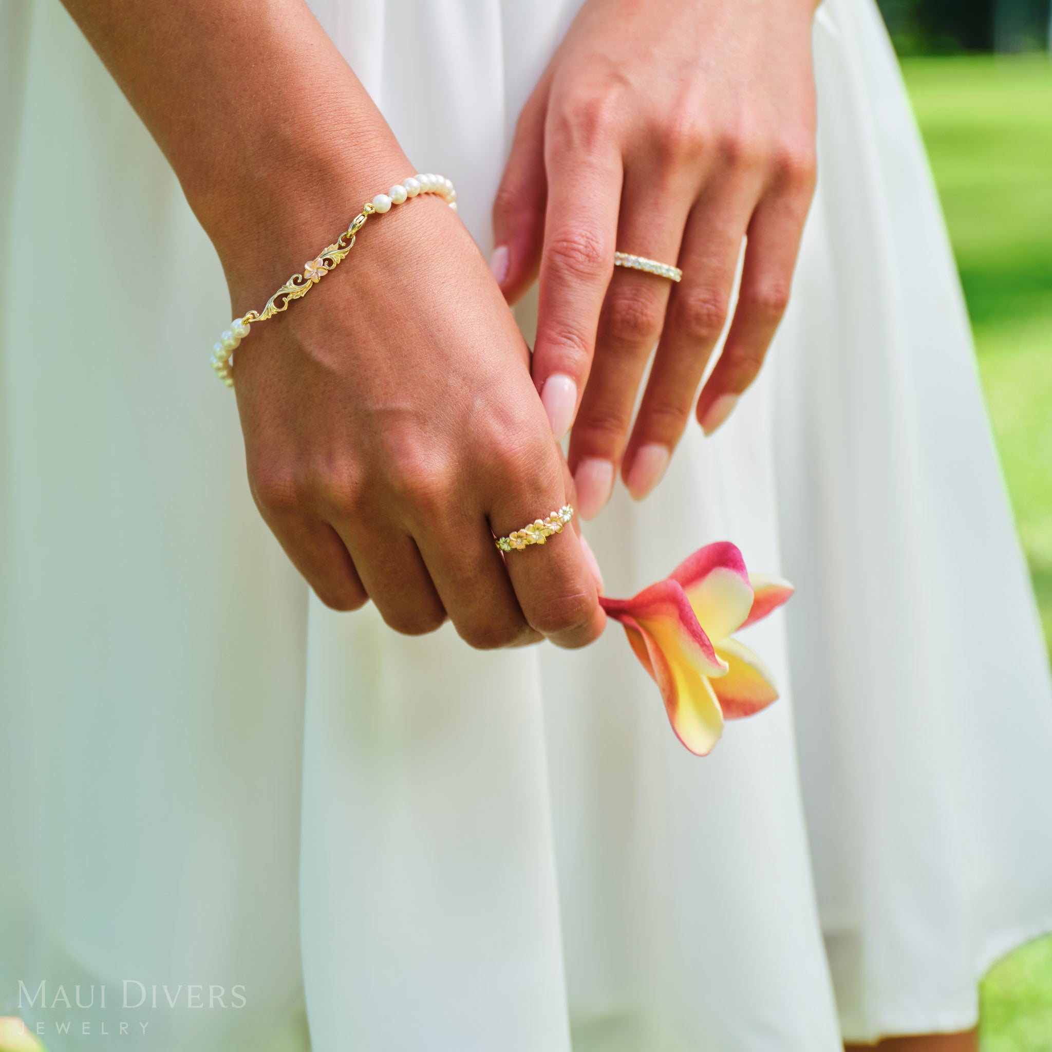 Close-up of a hand wearing a Hawaiian Heirloom Plumeria Freshwater White Pearl Bracelet in two tone gold with diamond and a Plumeria Tri Color Ring, holding a plumeria flower