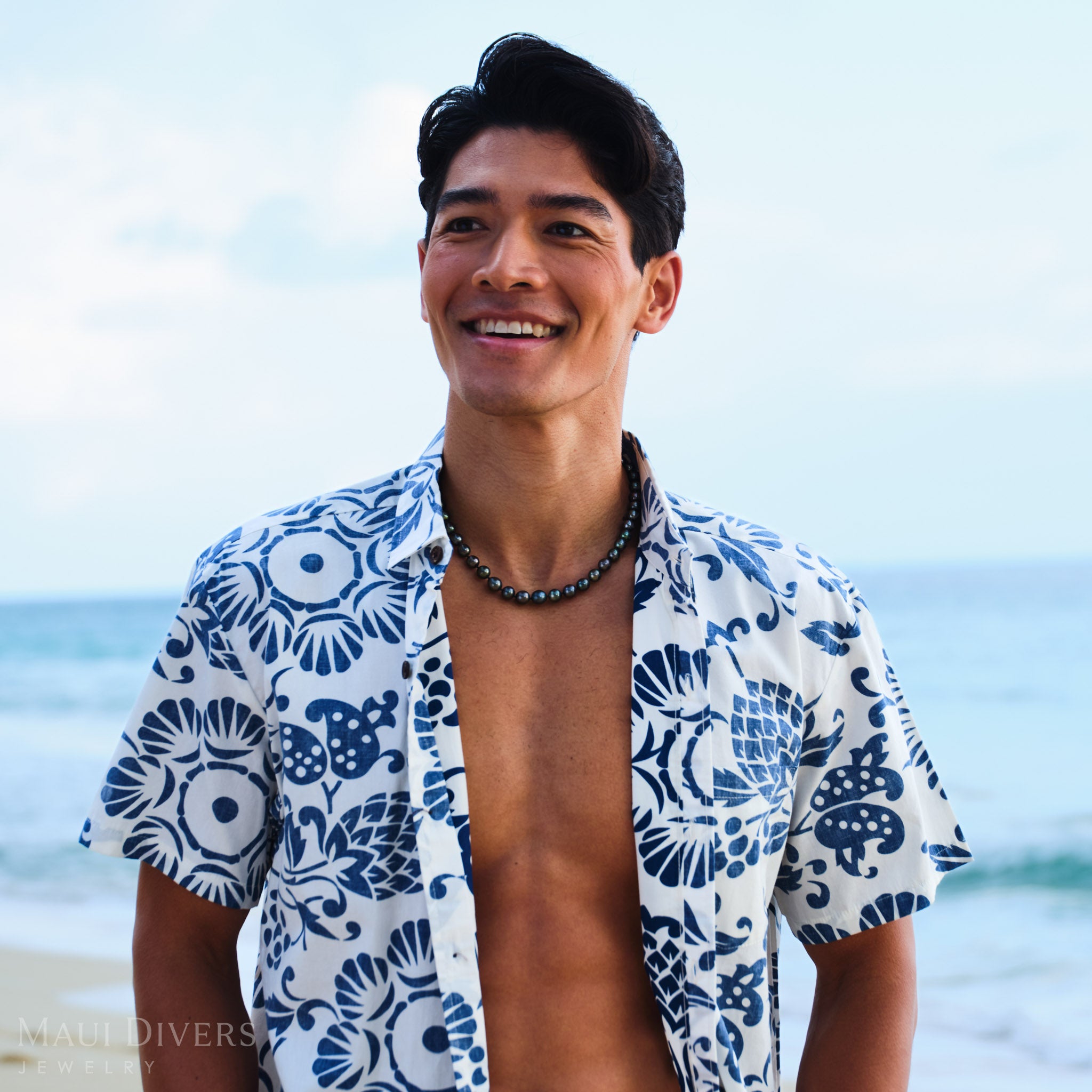 Man wearing a blue and white patterned shirt with a Tahitian black pearl strand, against a blurred beach background.