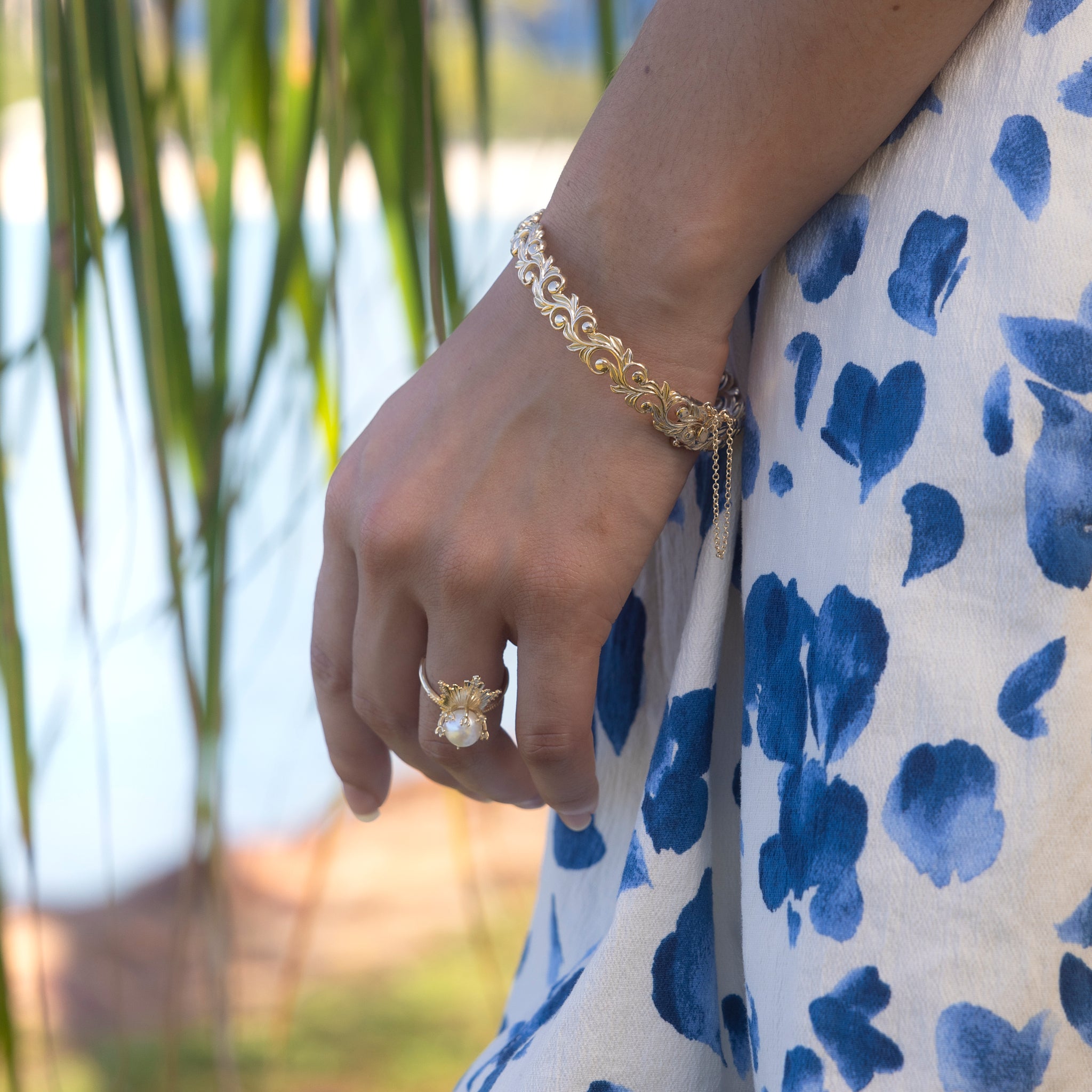 Close-up of a Living Heirloom Hinge Bracelet in 14k Yellow Gold on a wrist with a blurred palm and ocean background