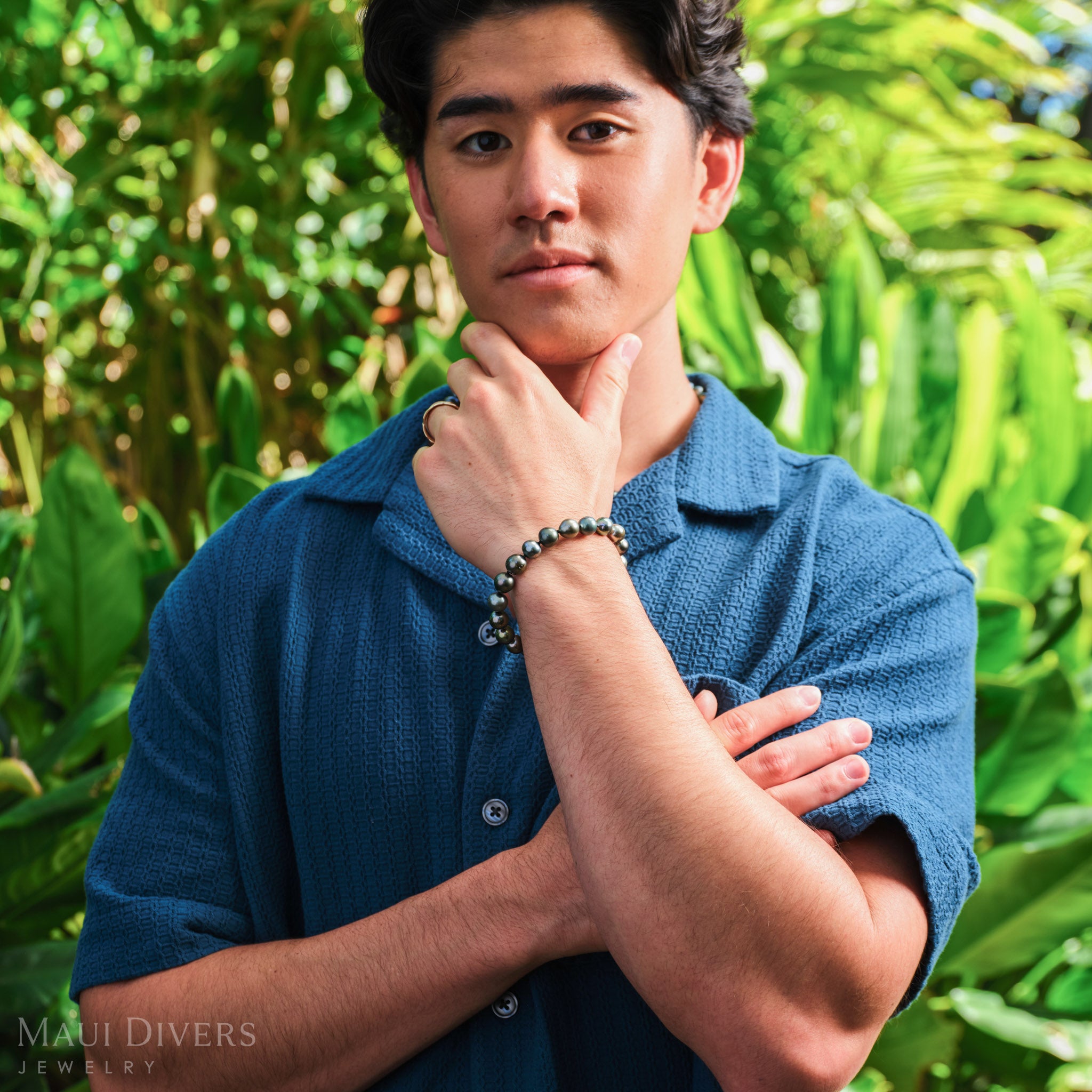 Man in a blue shirt wearing a Tahitian Black Pearl Bracelet in 14k yellow gold, against a blurred leafy green background