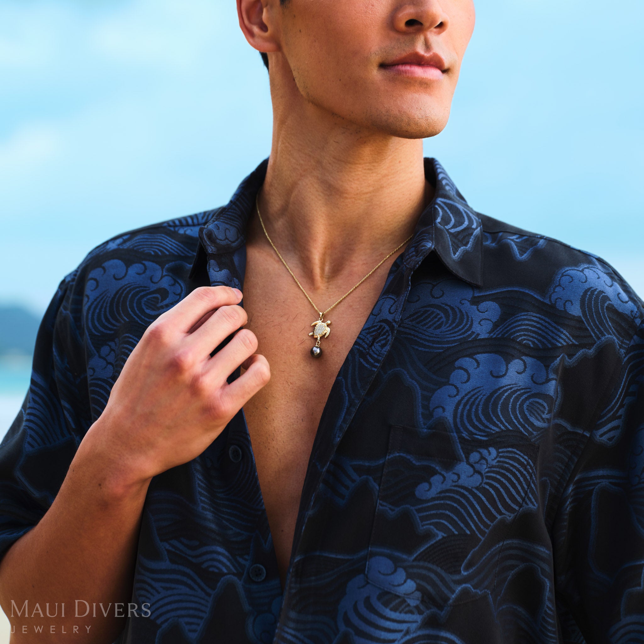 Man wearing a blue patterned shirt with a sea turtle Tahitian black pearl necklace against a blurred natural background