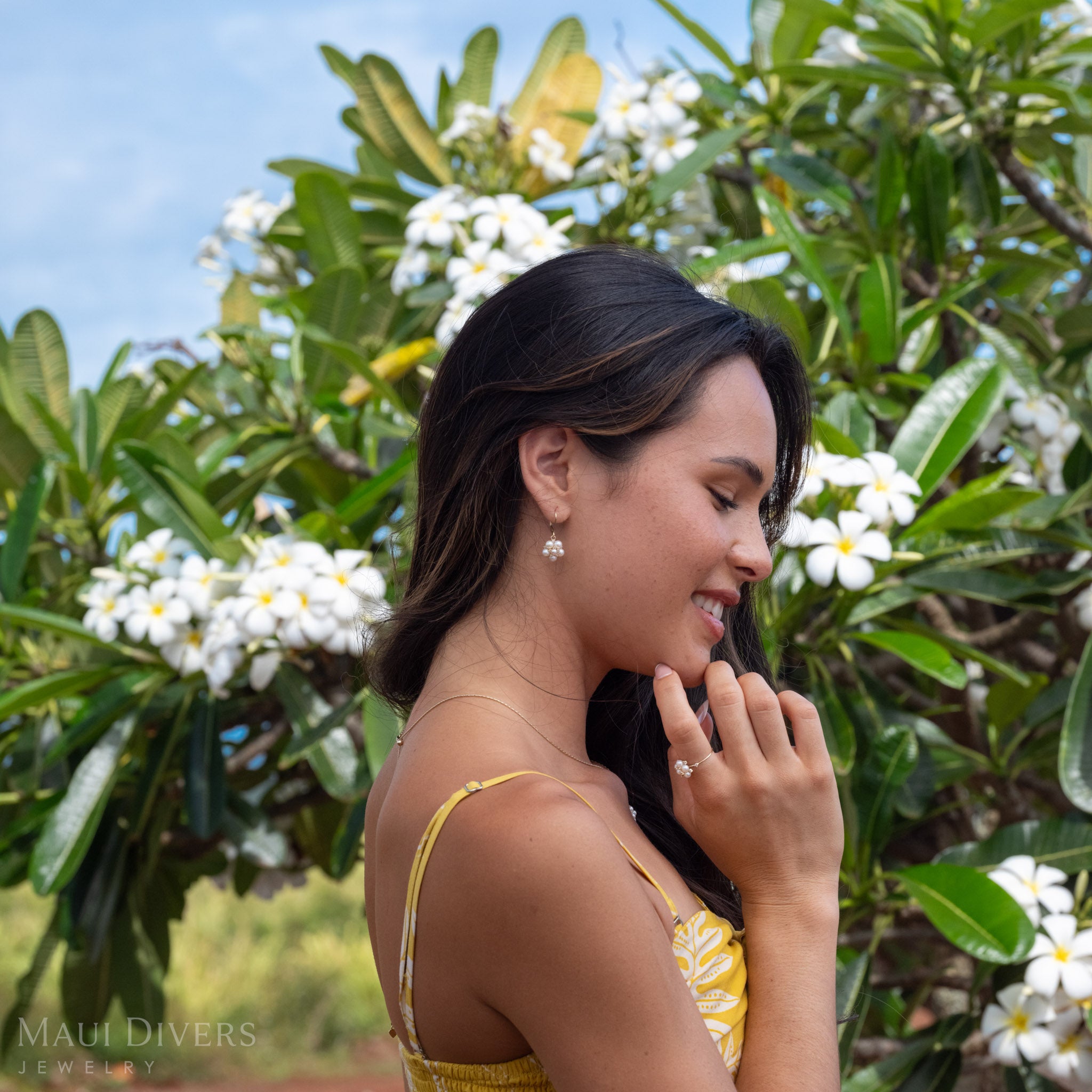 Side profile of a woman wearing Tiny Natural Freshwater White Pearls on a 14k Gold earring accented with Diamonds, her hand on her chin with the matching ring on her index finger against a blurred green and white plumeria background.