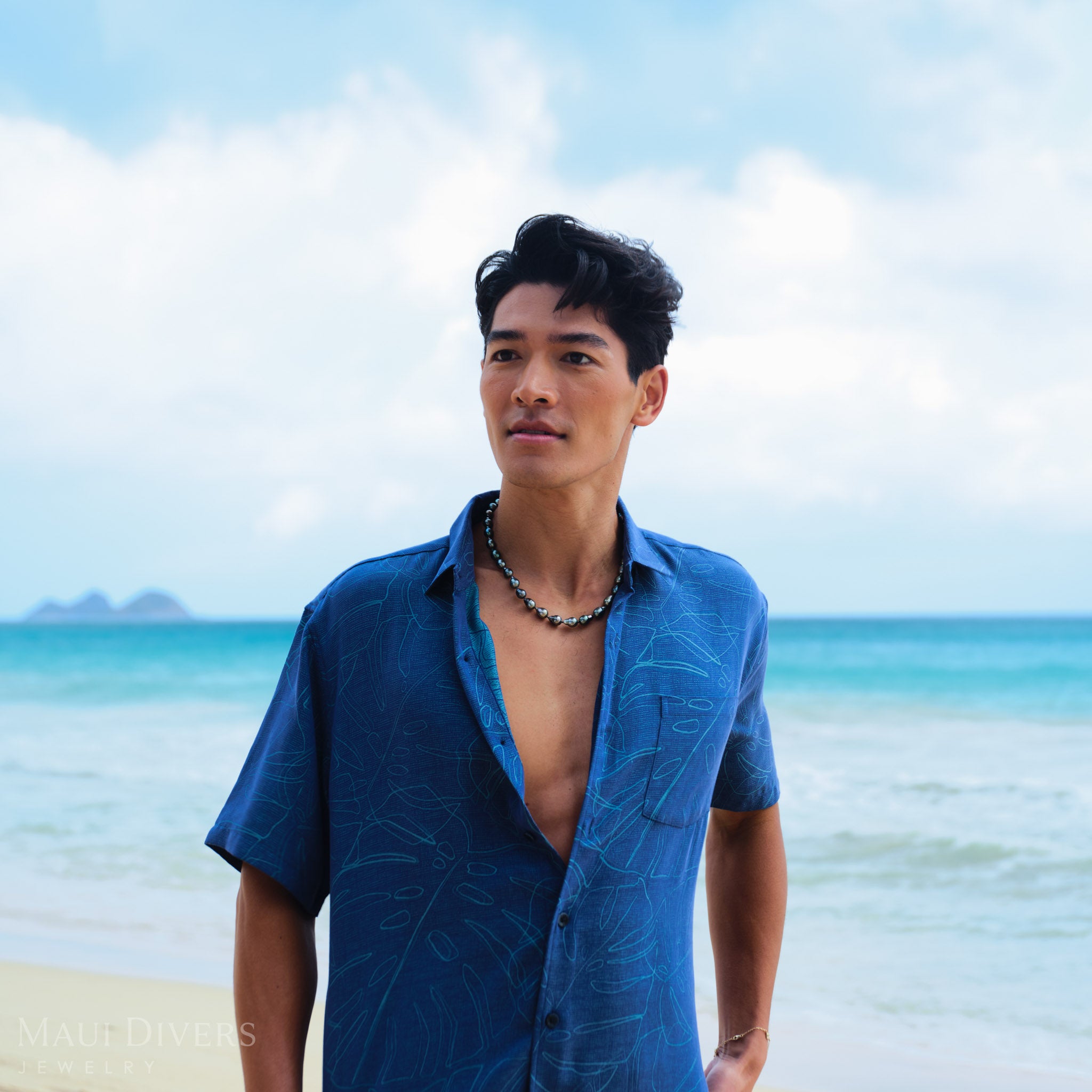 Man wearing a blue shirt and a Tahitian black pearl strand on a beach with clear blue water and sky.
