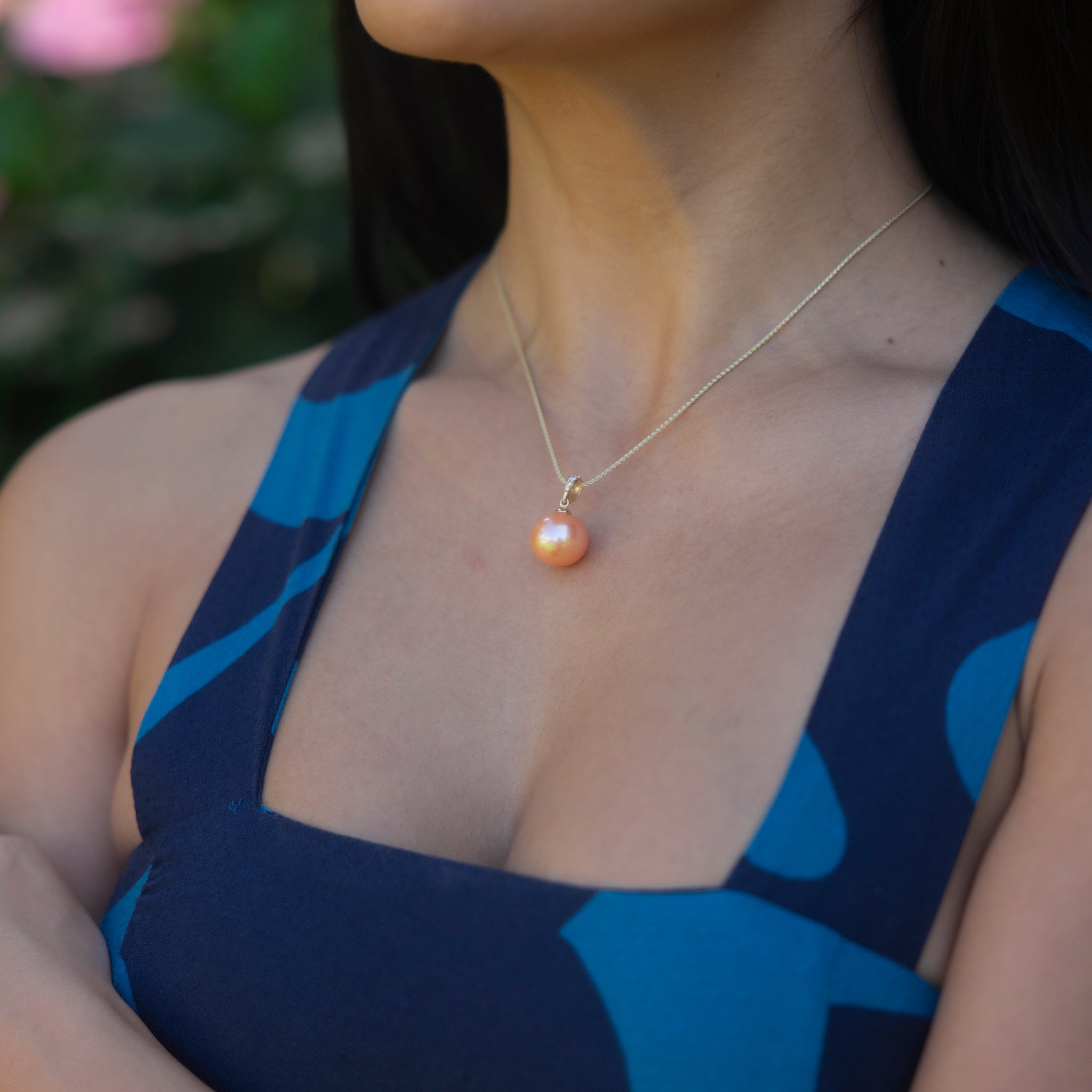 Person wearing a peach freshwater pearl pendant with diamond accents with blurry green background