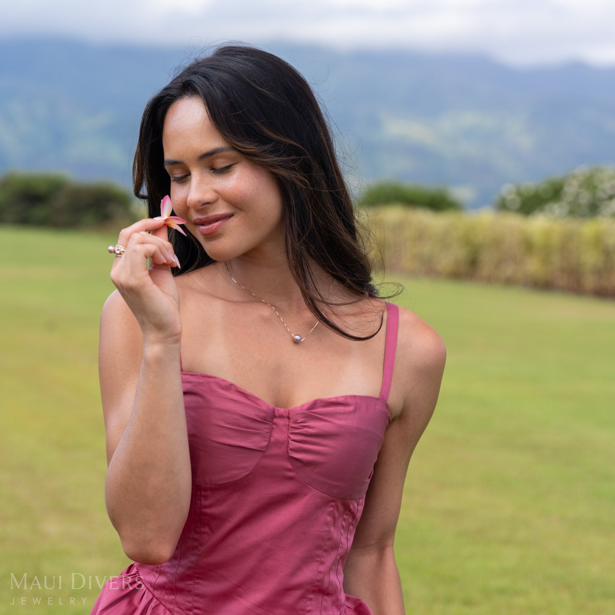 Smiling woman in a pink dress wearing a Freshwater Lilac Pearl Necklace in 14k yellow gold around her neck, smelling a plumeria flower in her hand, against a blurred open field background