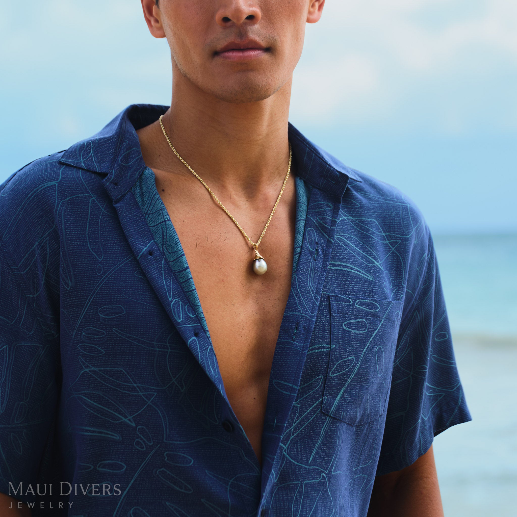 Man wearing a blue patterned shirt with a gold waterfall Tahitian black pearl pendant, set against a blurred beach background.