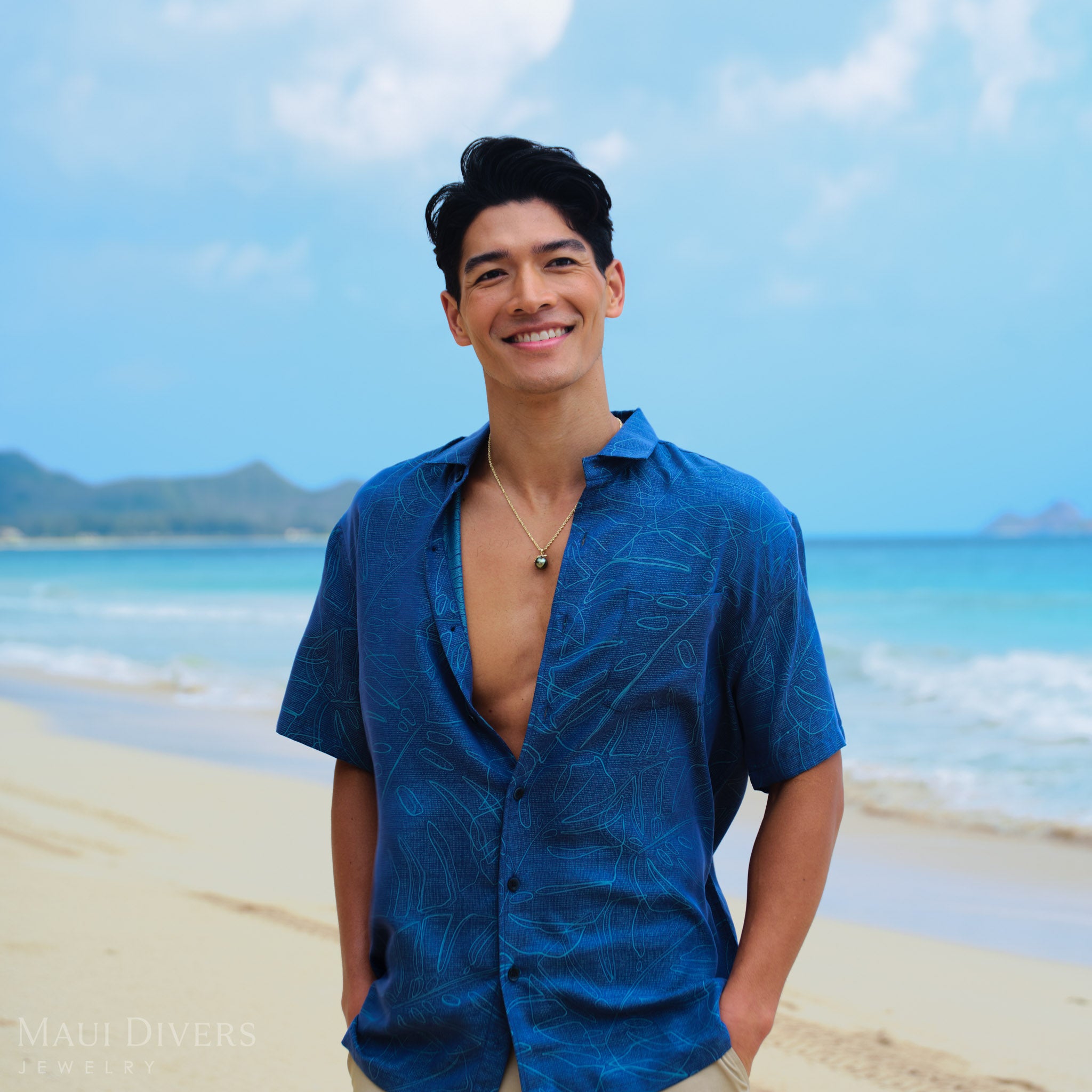 Man in a blue shirt and wearing a gold Tahitian black pearl pendant standing on a beach with clear blue water and sky.
