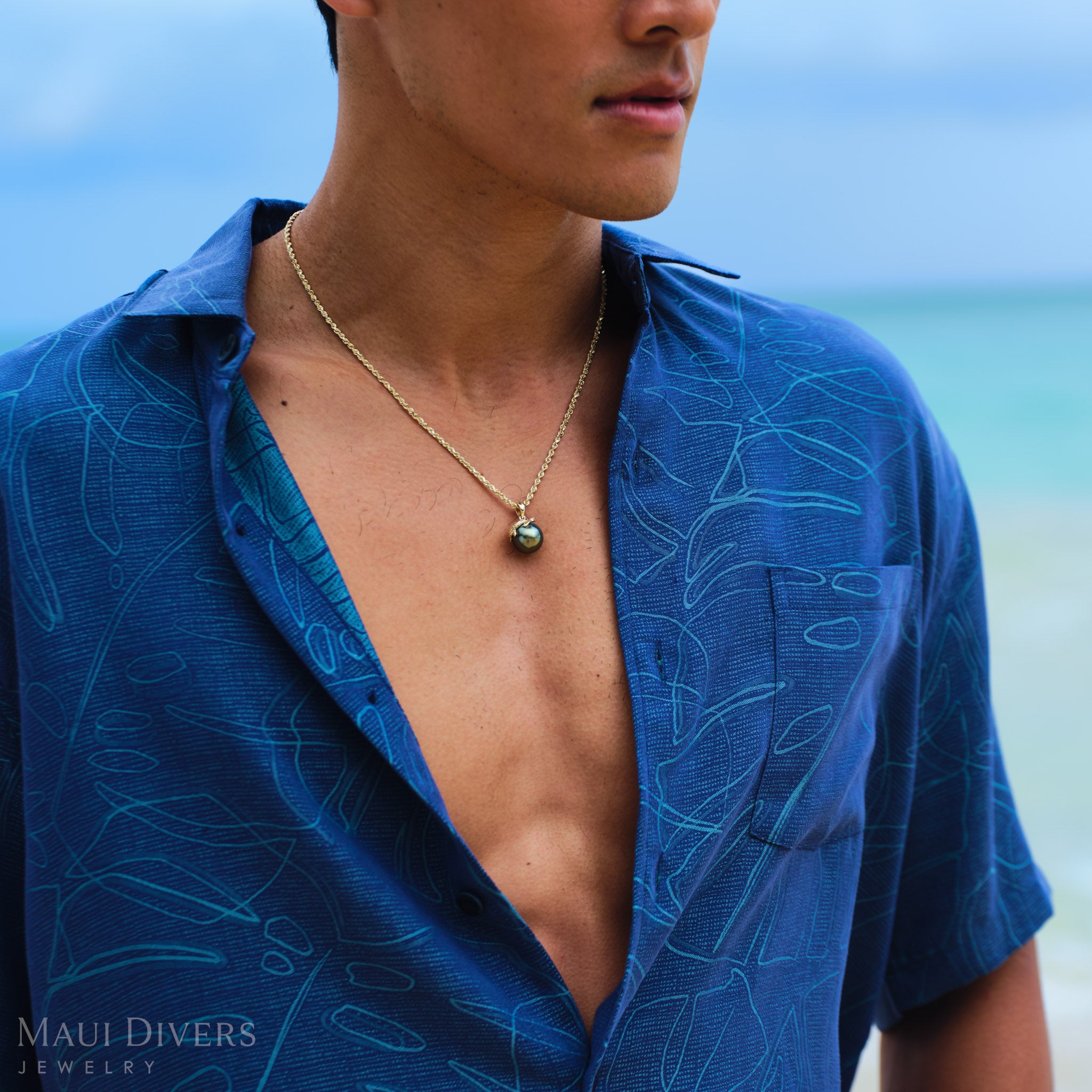 Man wearing a blue patterned shirt with a gold sea turtle Tahitian black pearl pendant on a beach