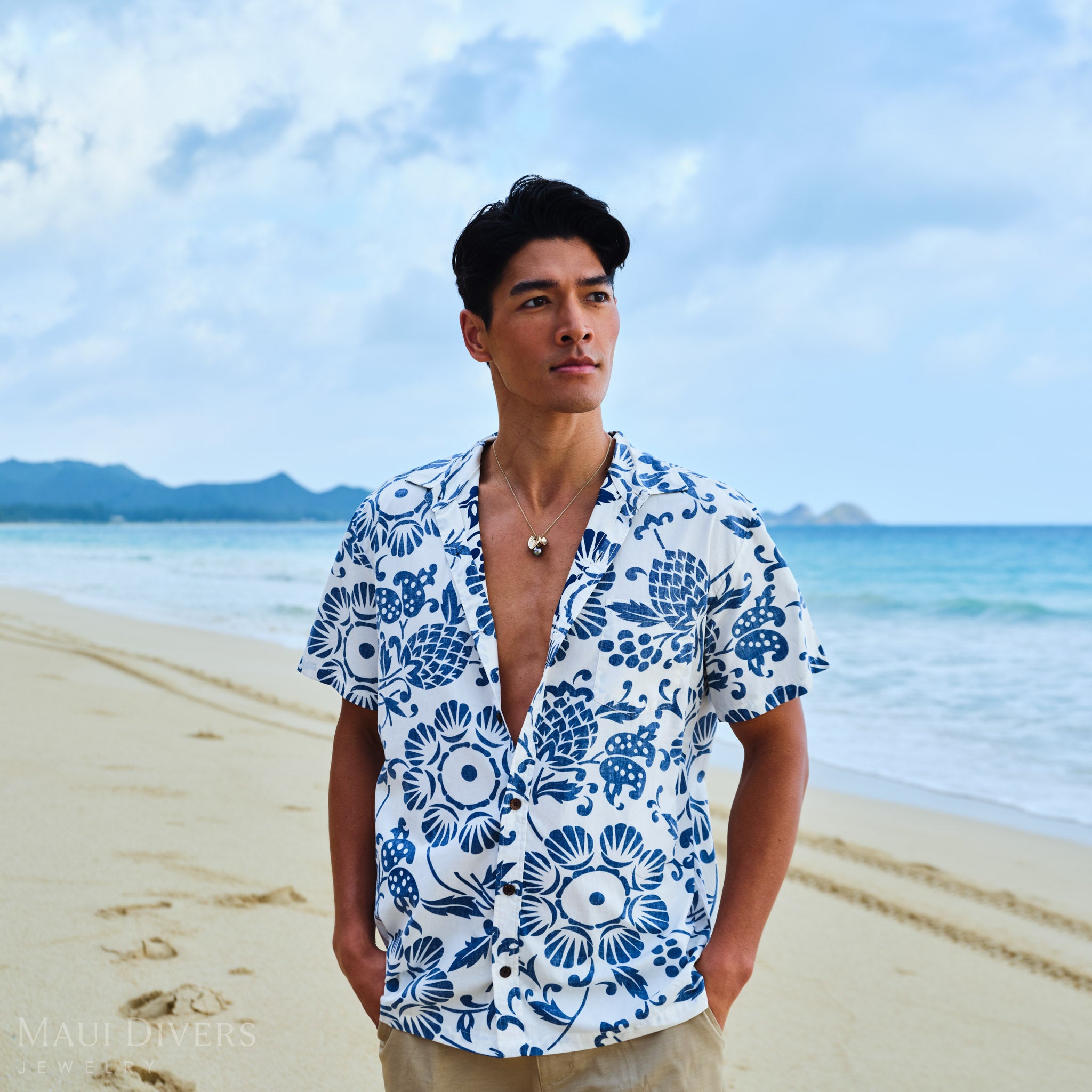Man wearing a blue and white patterned shirt with a Kalo Tahitian black pearl necklace, on a beach.