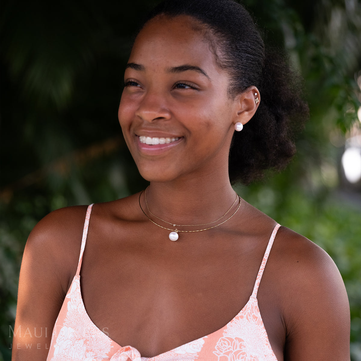 A smiling woman in a pastel colored top wearing a Freshwater White Pearl Pendant in 14k yellow gold with diamonds on an omega chain, layered with a tiny diamond heart necklace, a matching pair of Freshwater White Pearl Earrings in 14k yellow gold, against a blurred leafy green background