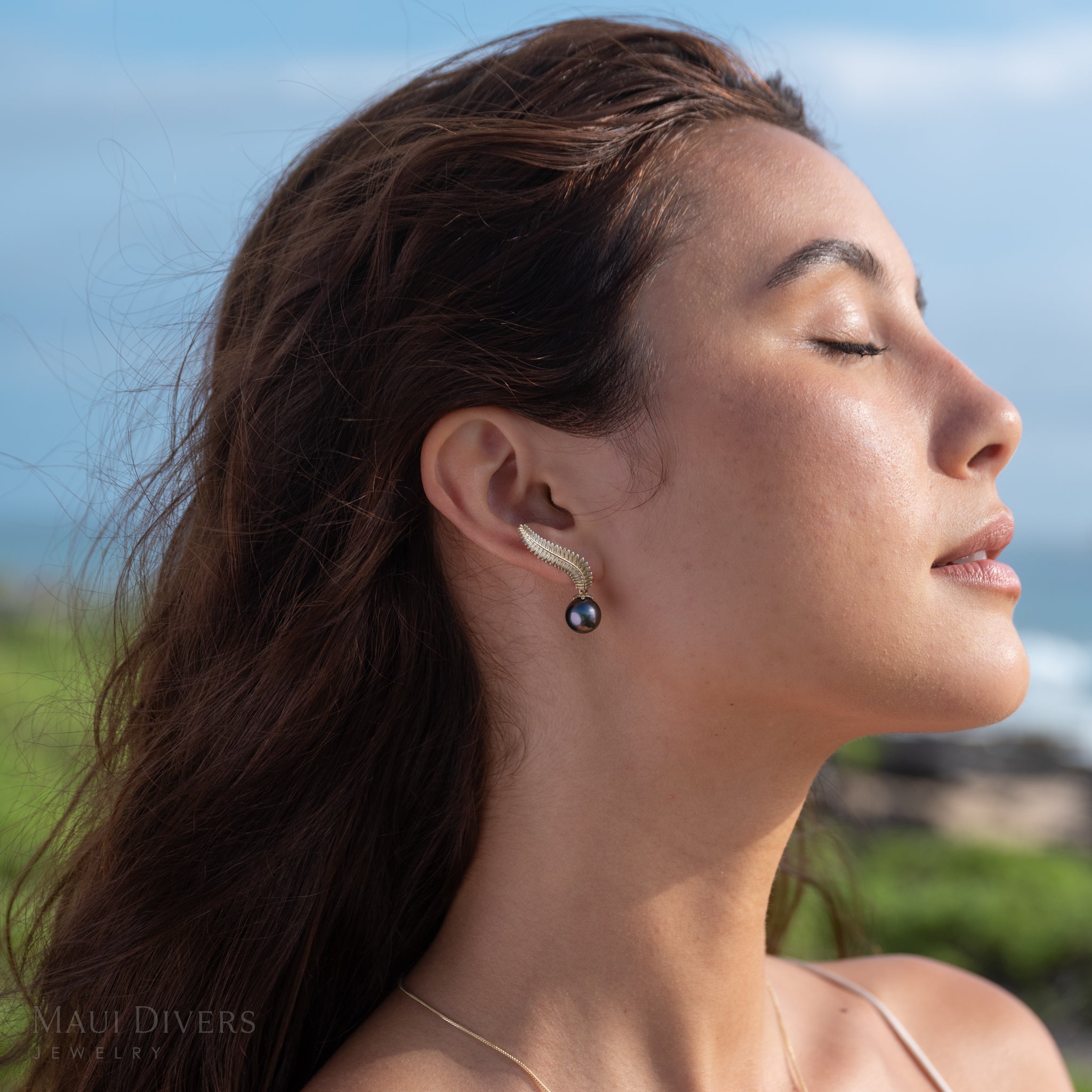 Close-up of a woman wearing 14k yellow gold Kupukupu fern Tahitian black pearl climber style earrings with a scenic beach background