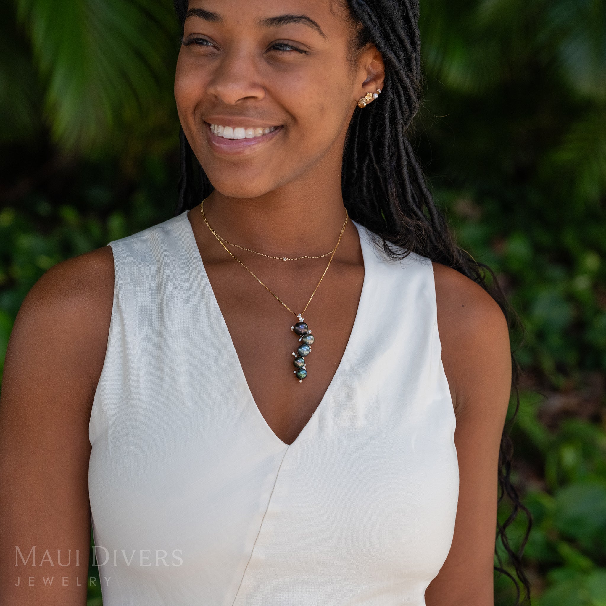 A smiling woman in a white top wearing a Bubbling Tahitian Black Pearl Pendant in 14k yellow gold with diamonds on a box chain, layered with a tiny diamond heart necklace, and an earring stack with an ʻOpihi earring, plumeria earring, white pearl earring, and diamond earring, against a blurred leafy green background