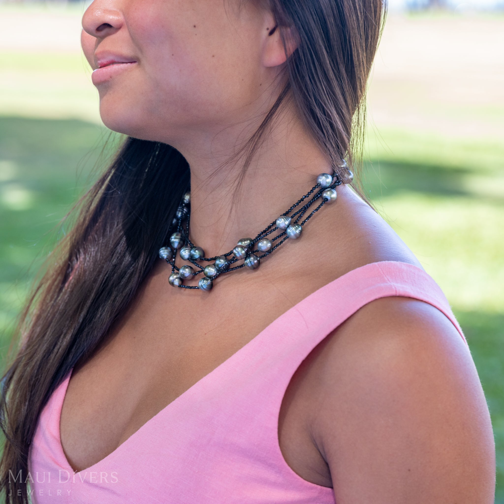 A close-up sideview of a smiling woman in a light pink top wearing a Tahitian Black Pearl and Black Spinel Layered Necklace with a 14k yellow gold clasp, against a blurred green grass background