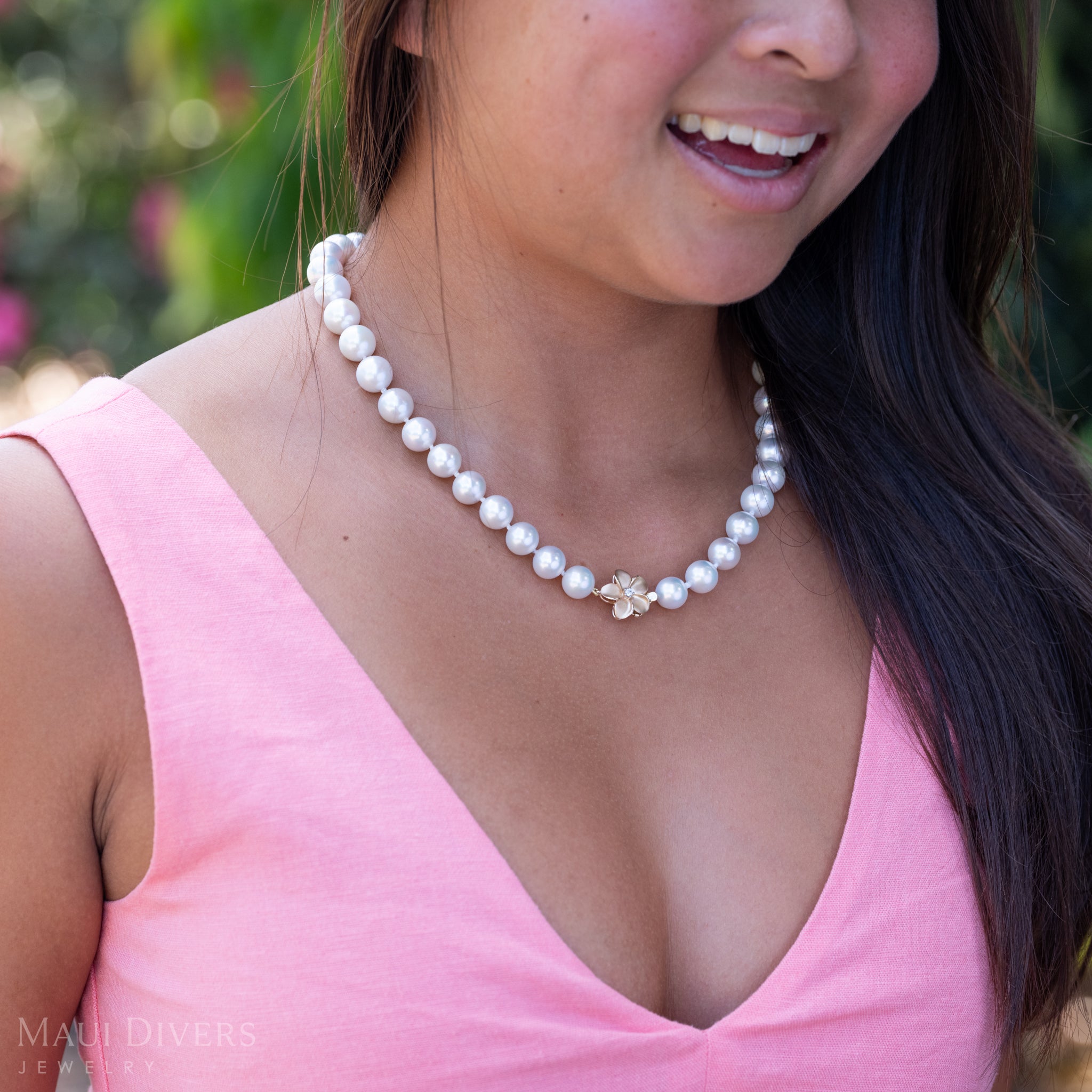 A close-up of a woman in a light pink top wearing a Freshwater White Pearl Strand with Diamond Plumeria Clasp in 14k Yellow Gold, against a blurred background