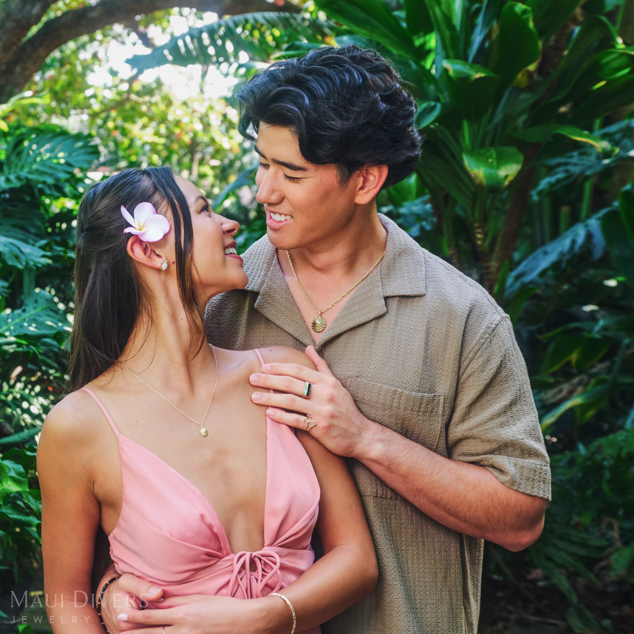 Couple embracing wearing matching ʻOpihi pendants in 14k yellow gold with diamonds, against a tropical forest background