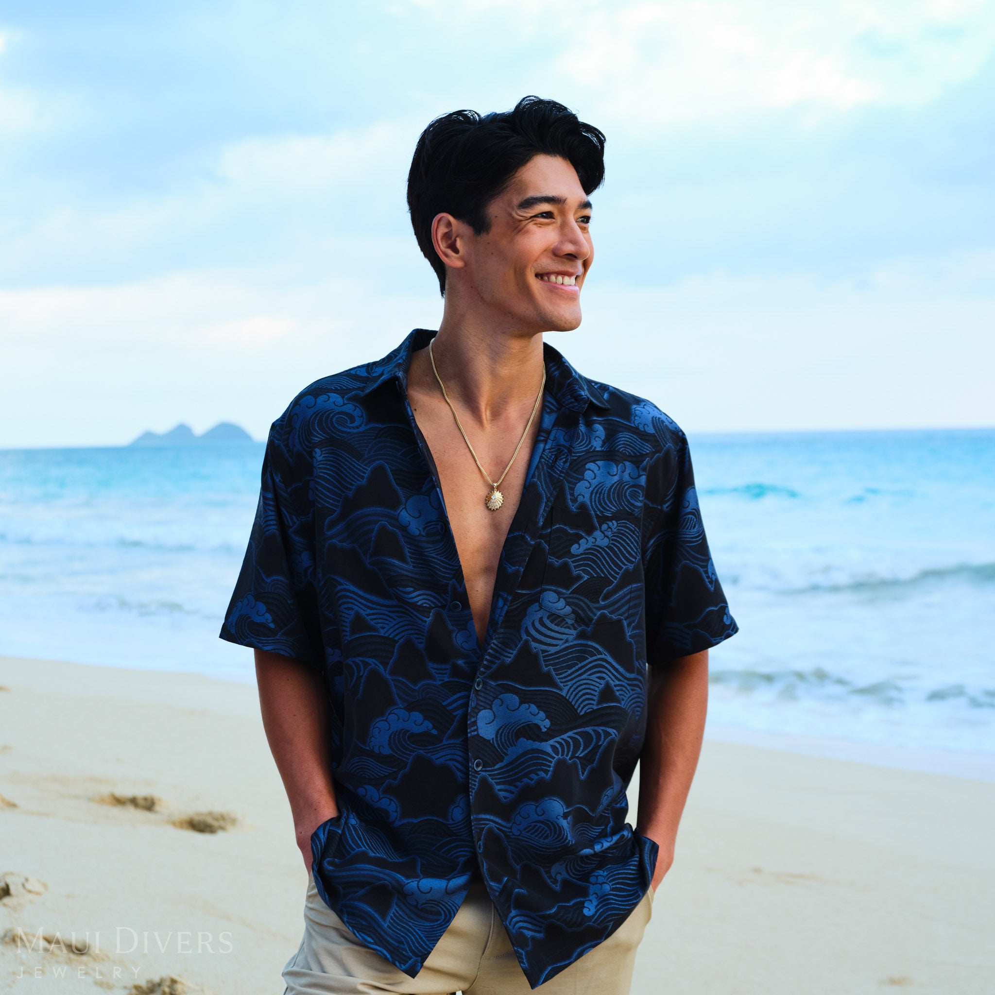 Man wearing a blue patterned shirt and an ʻopihi pendant on a beach with ocean and sky in the background