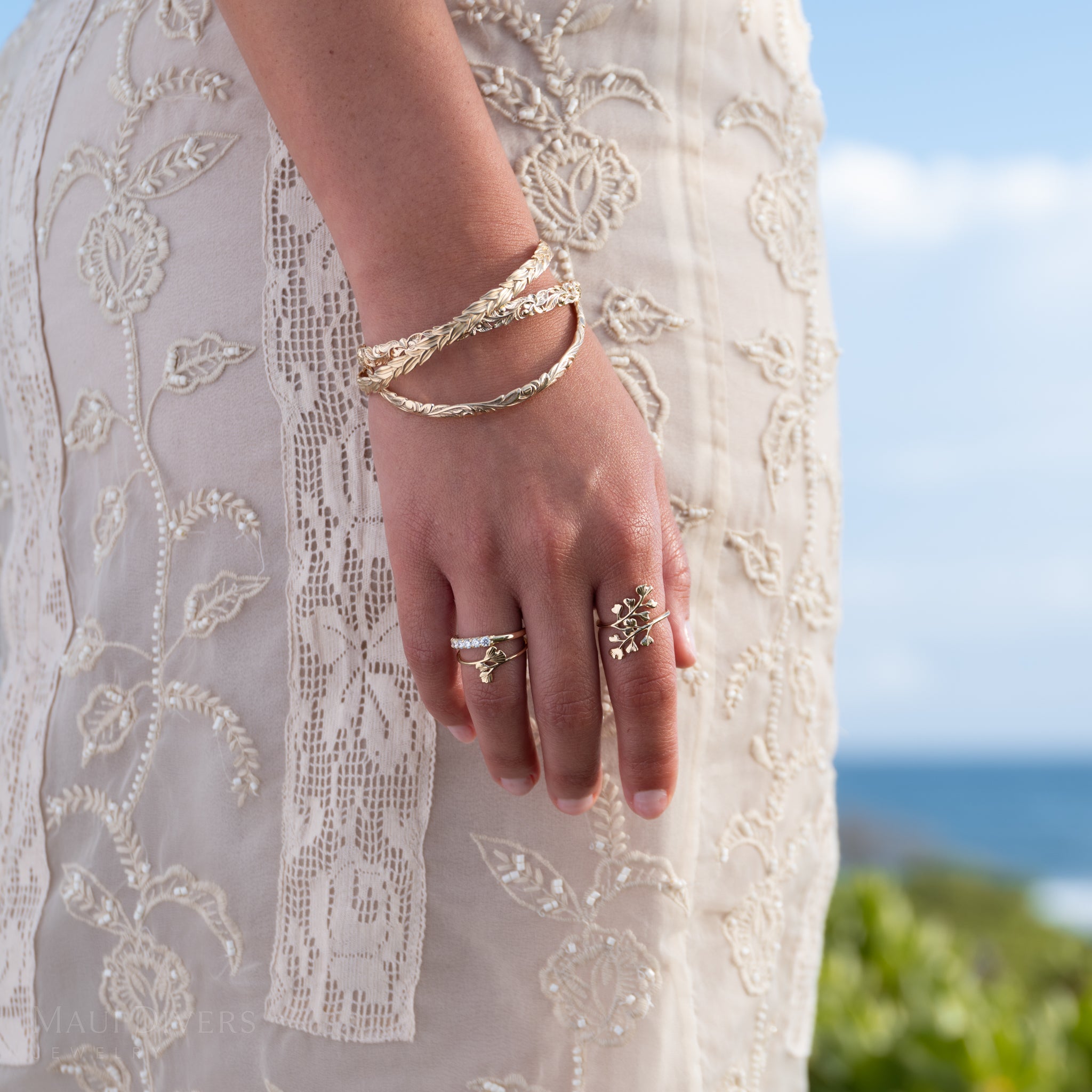 A close-up of a person's hand wearing multiple 14k yellow gold Maiden Hair rings and stacked 14k yellow gold Hawaiian Heirloom bracelets, styled with a cream lace dress against a blurred outdoor background of blue sky and ocean