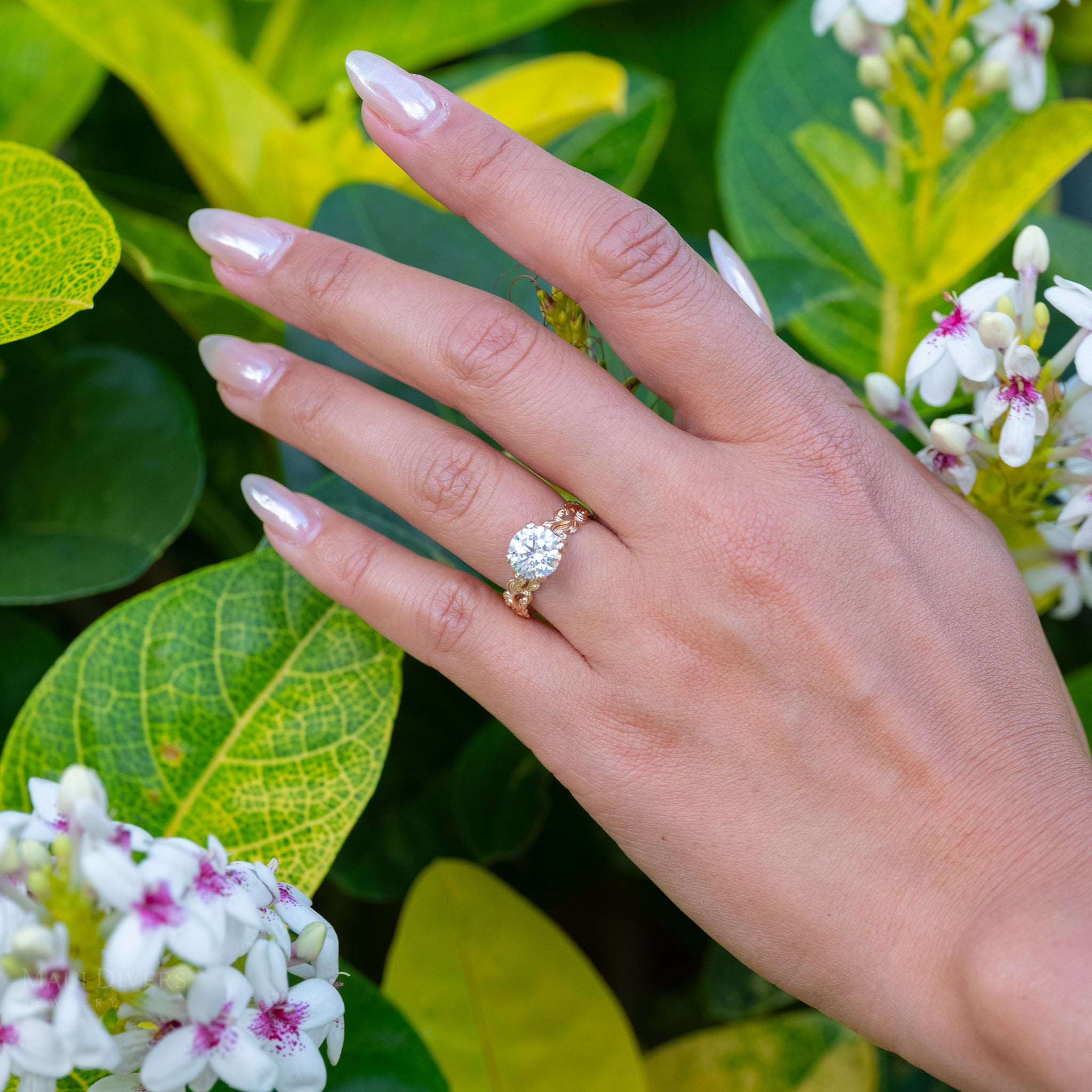 Hand wearing a solitaire lab-grown diamond engagement ring with green foliage and flowers in the background