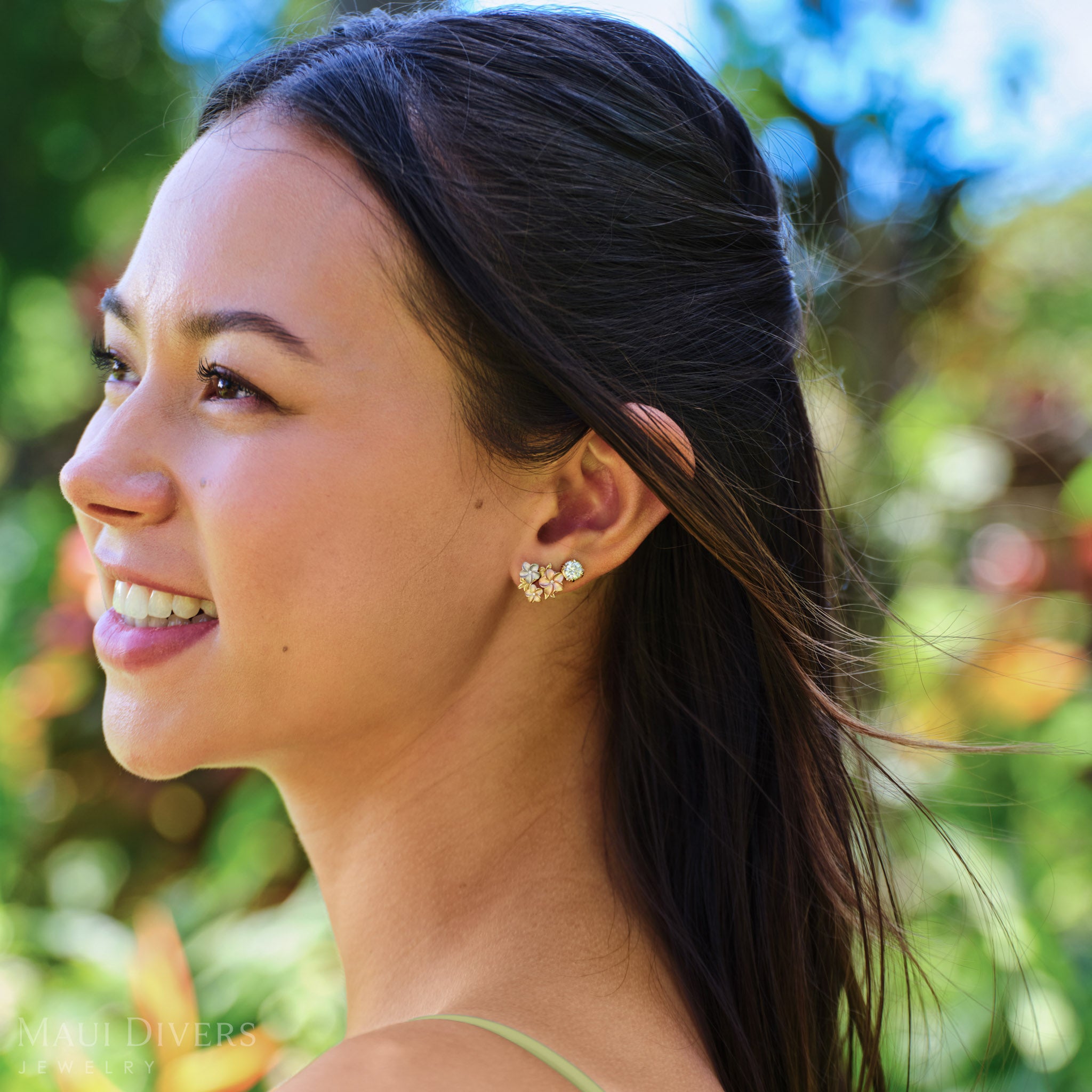 Side view of a smiling woman wearing a Hidden Hawaiʻi Maile Earring in 14k Yellow Gold with Lab Grown Diamond and a Plumeria Earring in Tri Color Gold with Diamonds on her left ear