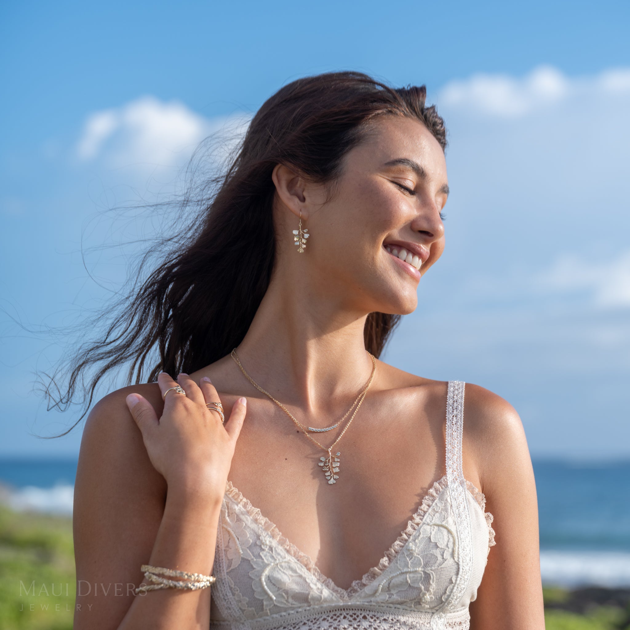 A woman outdoors by the ocean wearing a Maiden Hair Pendant and Earring, a diamond lavalier necklace, paired with a white lace top.