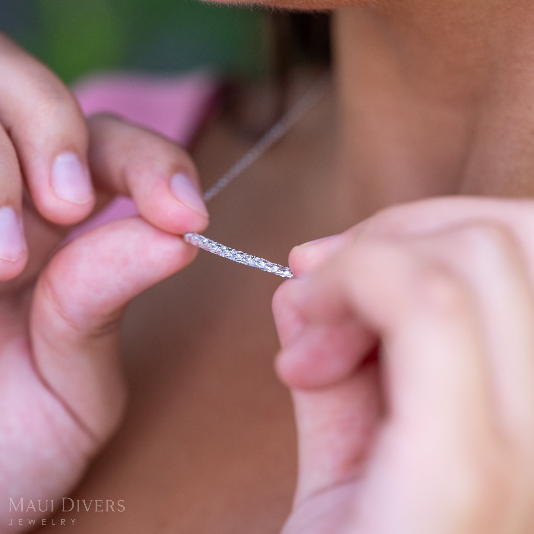 A close-up of a woman in a light pink top showing the design of an Adjustable Hidden Hawaiʻi Maile Lab Grown Diamond Necklace in 14k White Gold, against a blurred leafy background