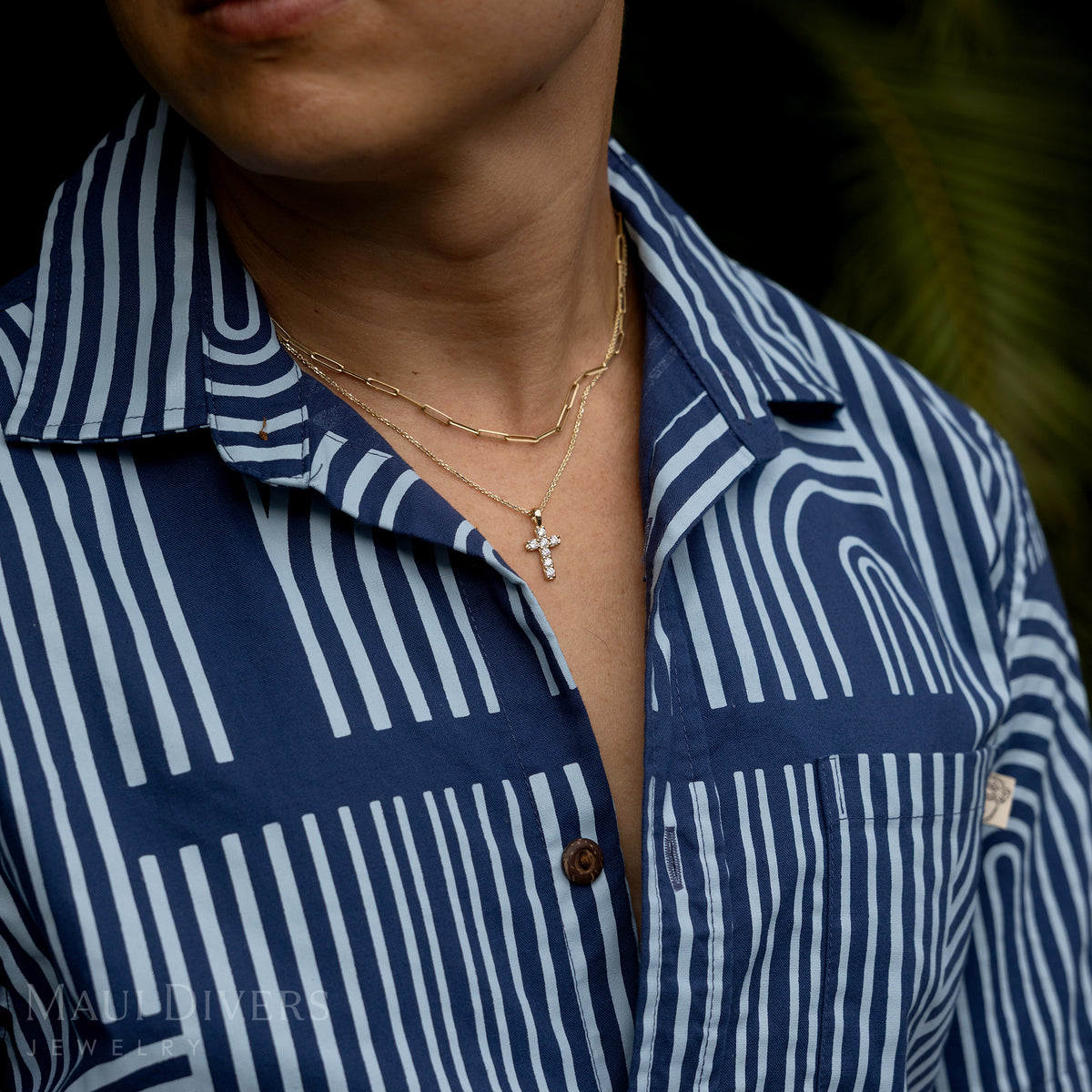 A close-up of a guy in a two-toned blue patterned shirt wearing a Hidden Hawaiʻi Maile Lab Grown Diamond Cross Pendant in 14k yellow gold around his neck, layered with a paperclip chain against a blurred green leaf background