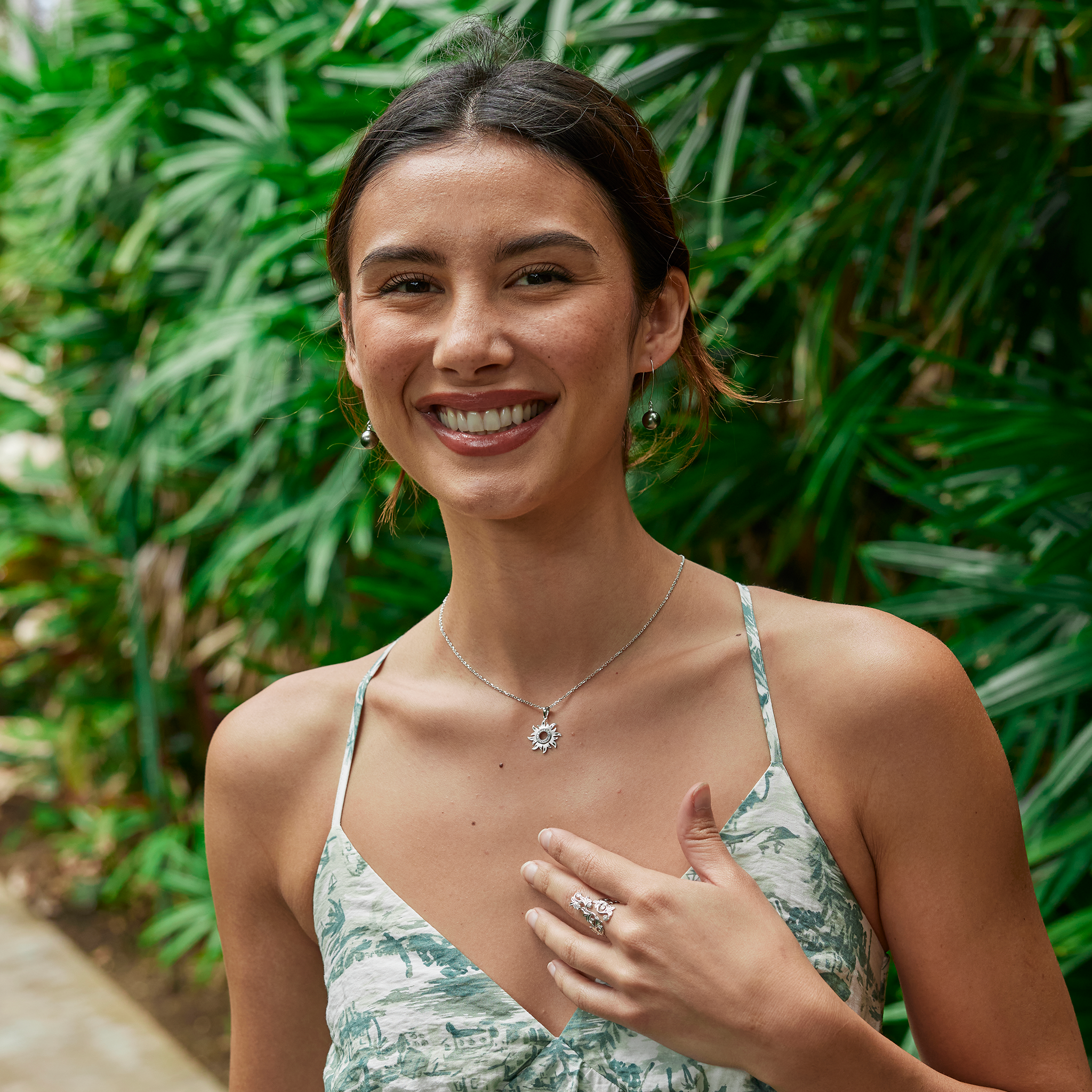Smiling woman wearing white gold sun pendant and tahitian pearl earrings with green foliage in background