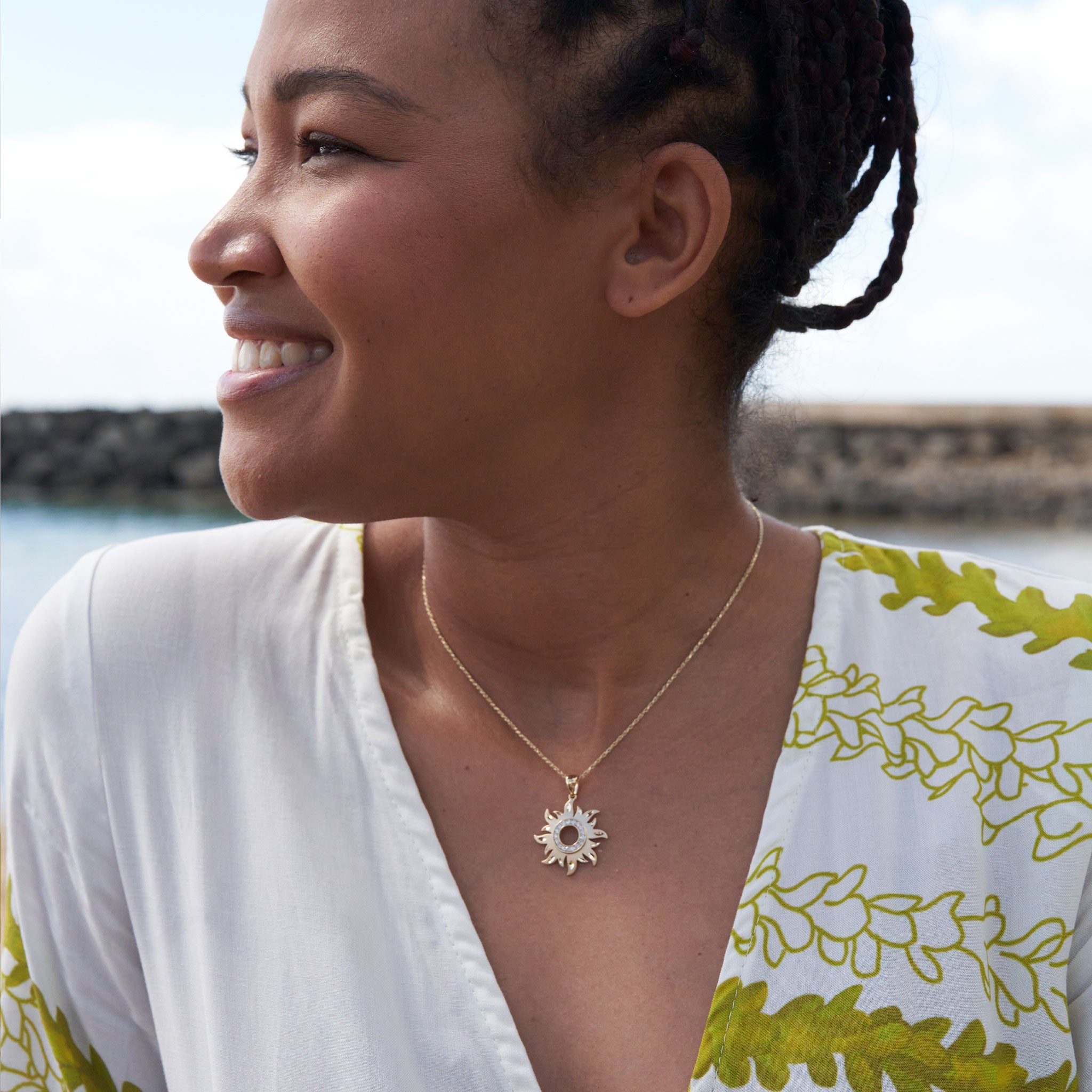 Smiling woman wearing a gold sun pendant with diamonds in front of water and a bright sky