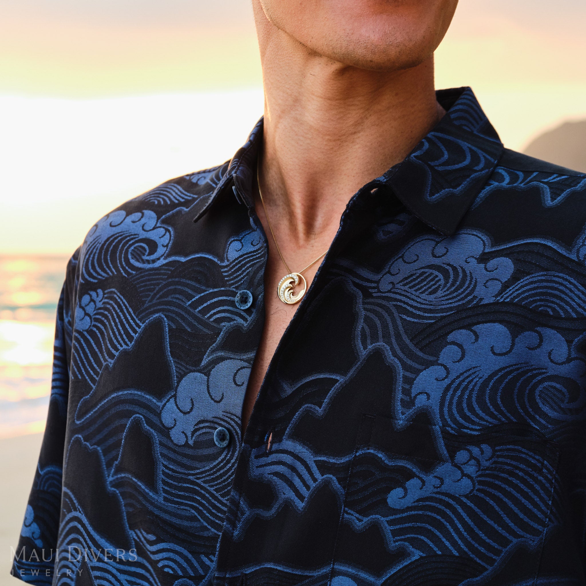 Man wearing a dark blue patterned shirt with a gold diamond wave pendant with a blurred sunrise background