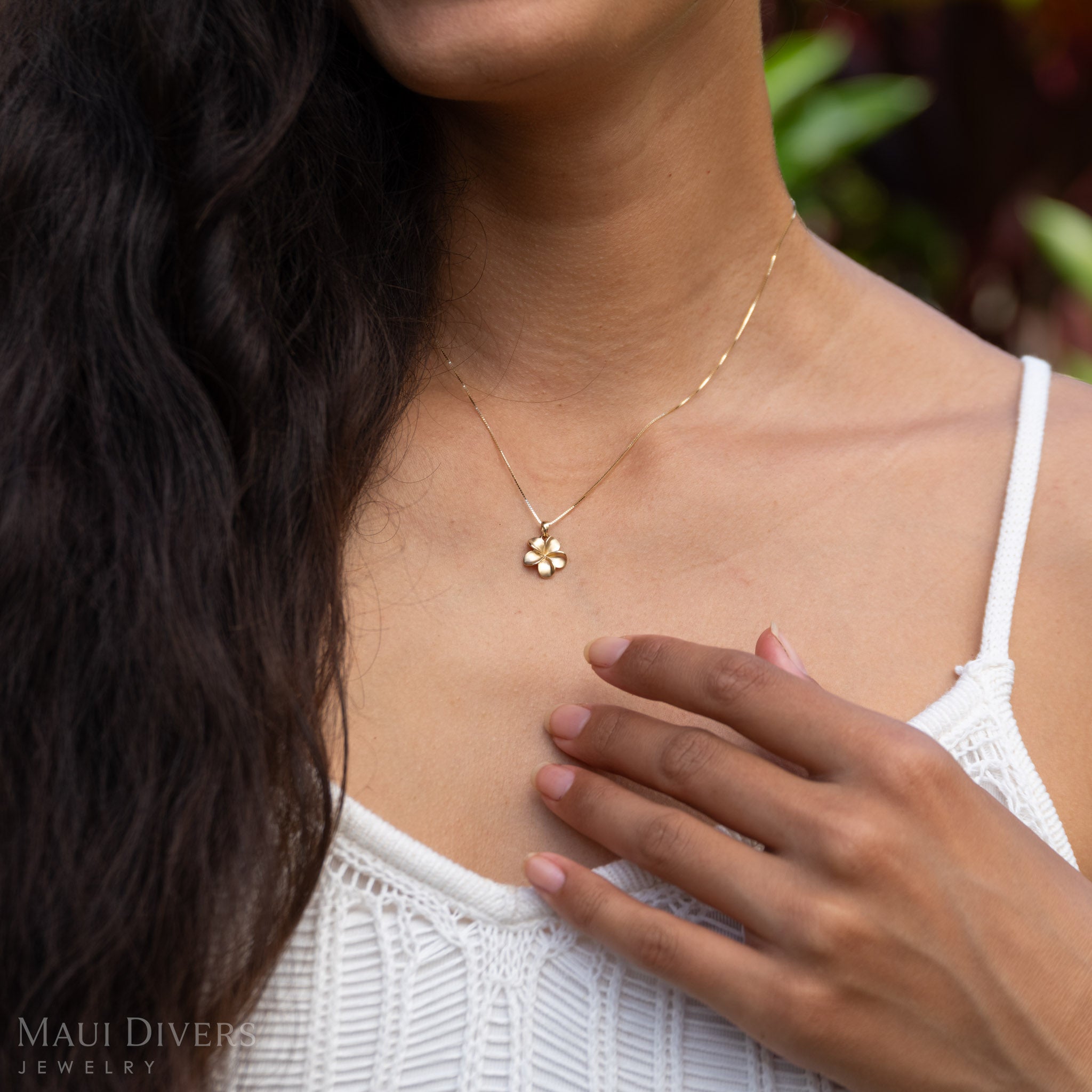 Woman wearing a gold necklace with a small plumeria pendant, outdoors.