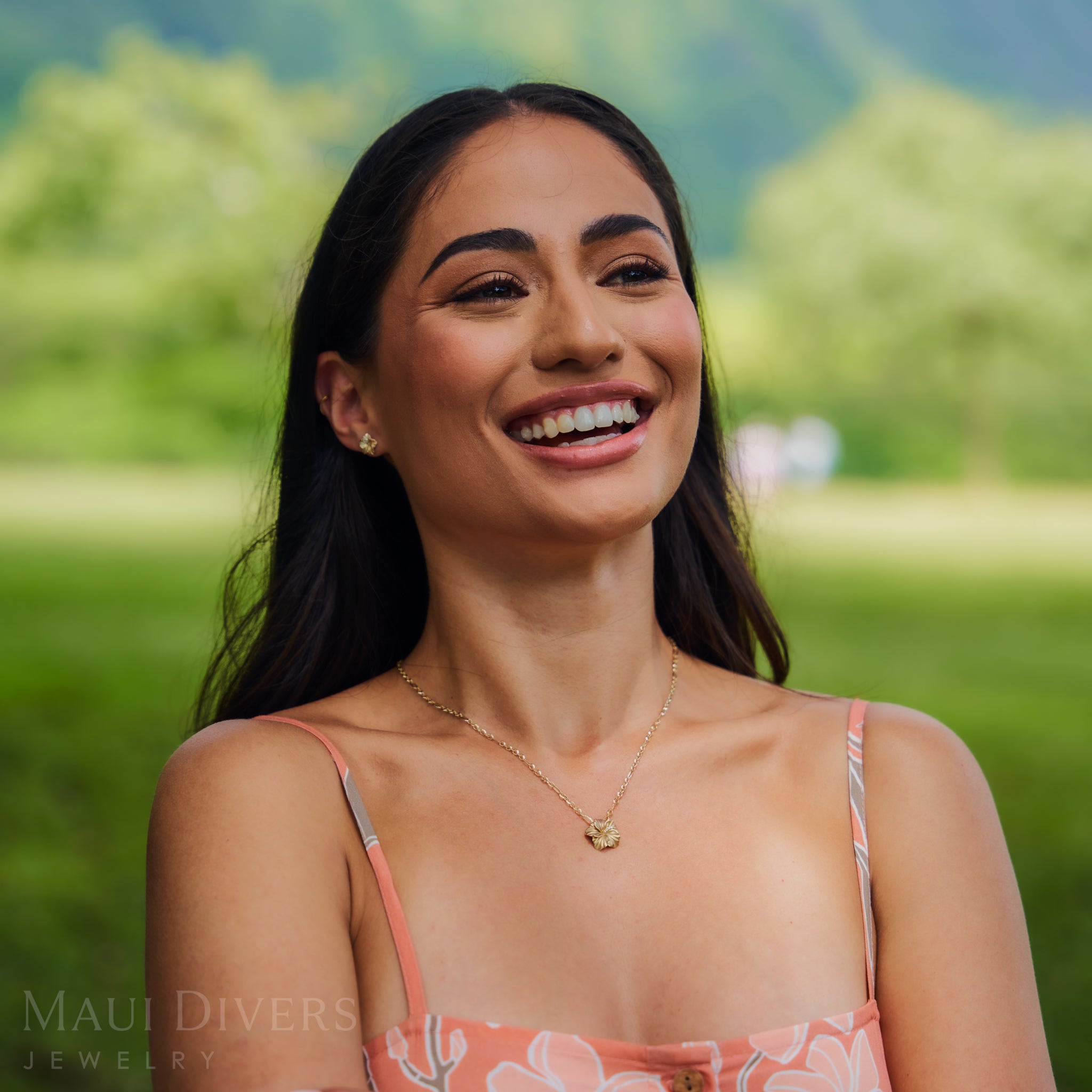 Woman smiling outdoors wearing a diamond hibiscus pendant with a blurred green background