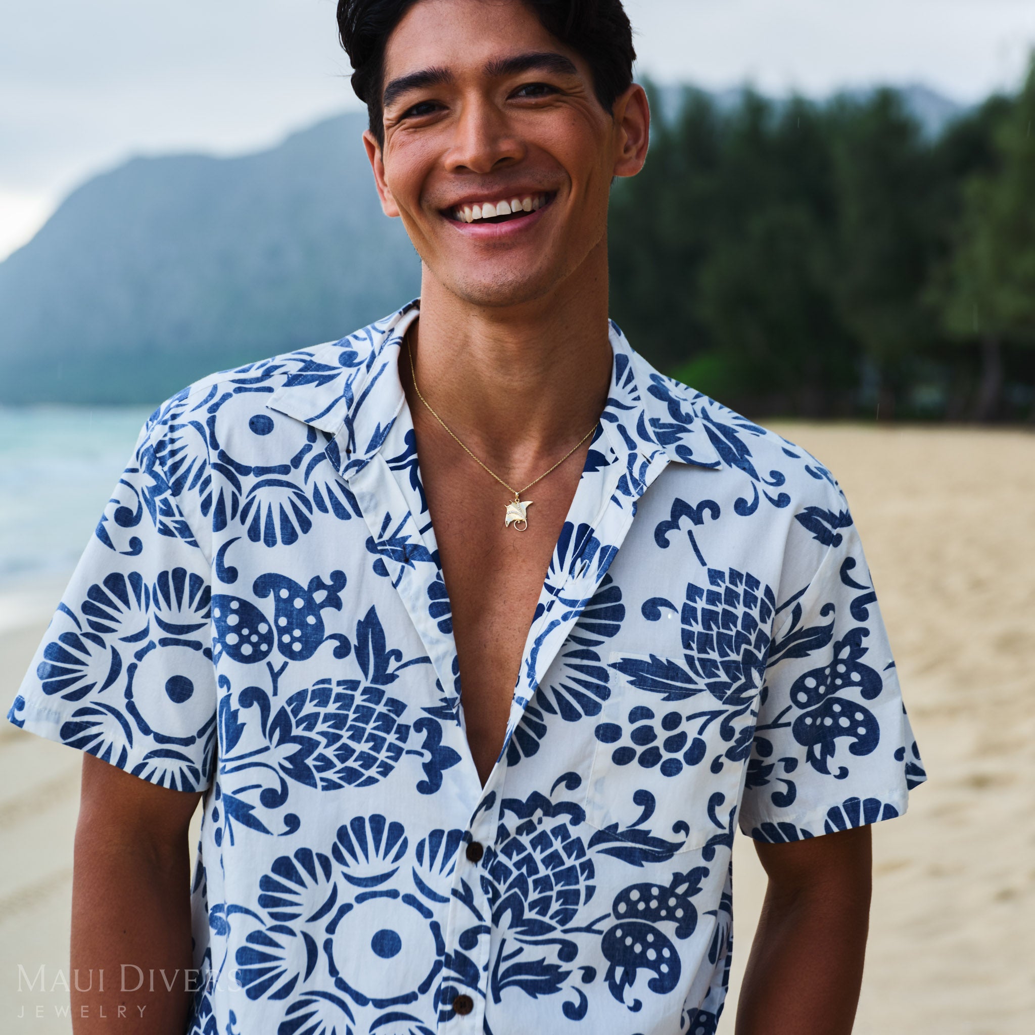 Man wearing a blue and white patterned shirt and a manta ray pendant on a rope chain on a beach.