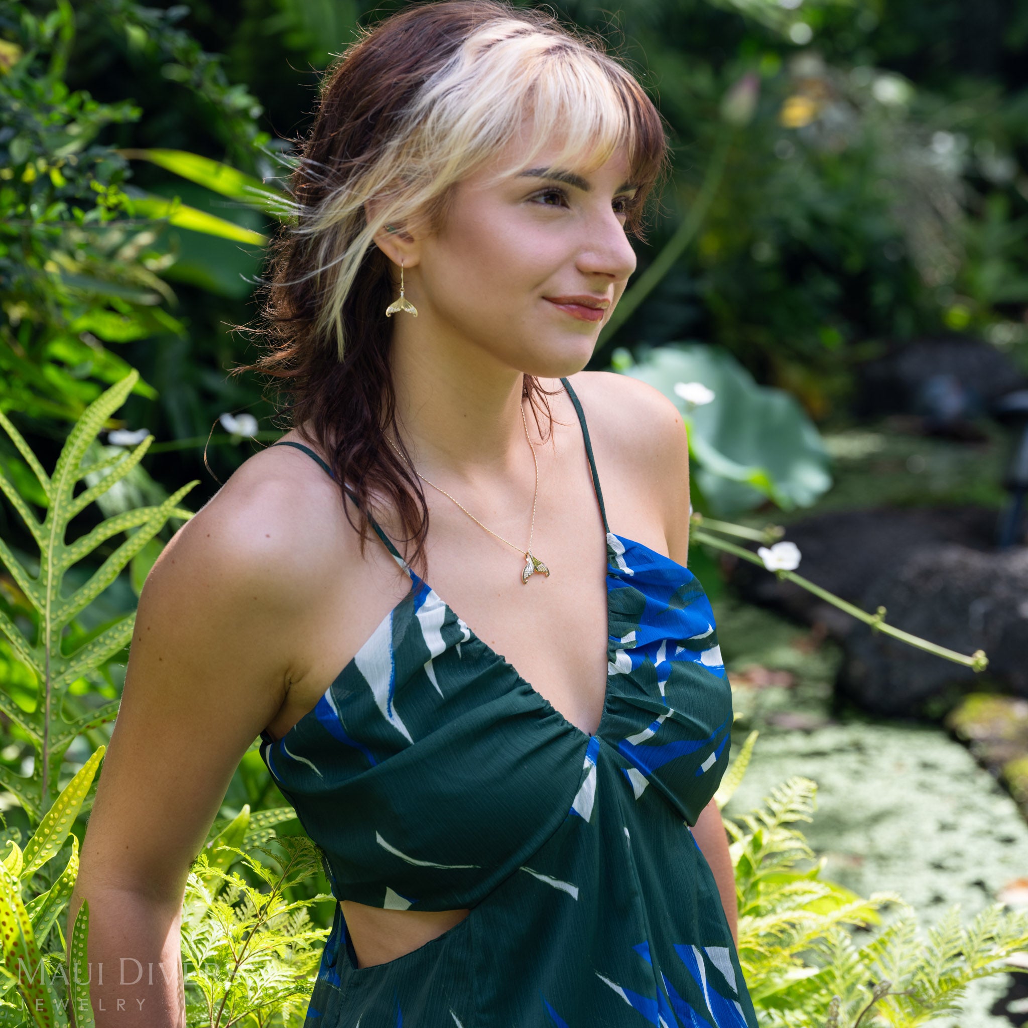 A woman wearing a dangling diamond mermaid tail leverback earring and matching pendant in 14k yellow gold and a dark green and blue patterned halter dress with a side cutout, standing in a lush green outdoor garden setting.