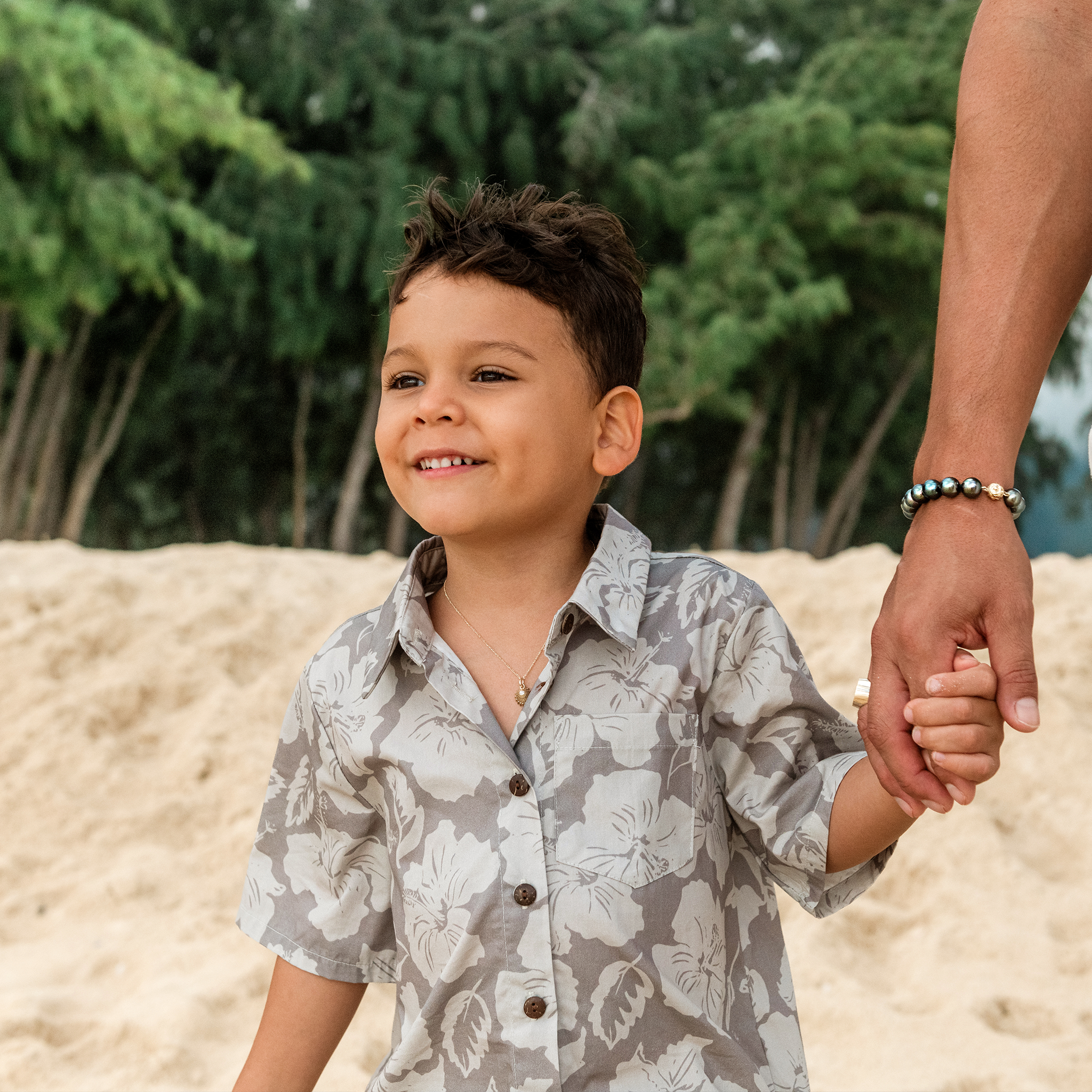 Boy at the beach wearing a gold necklace with an ʻOpihi diamond pendant holding a manʻs hand