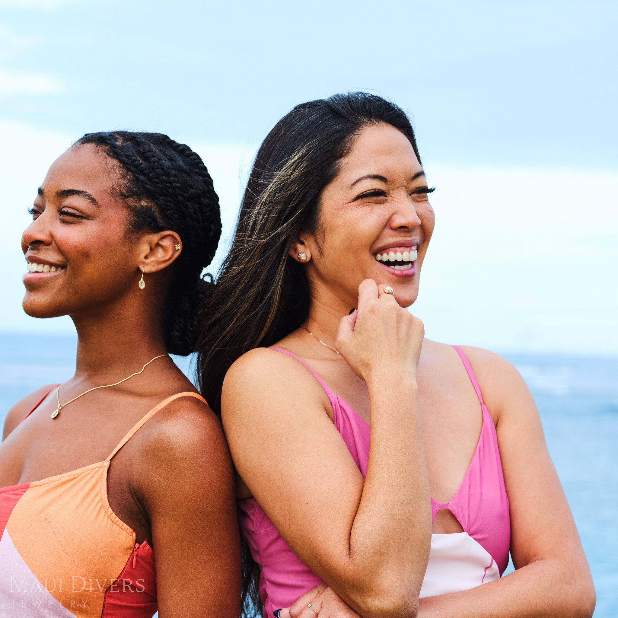 Two women laughing and smiling, one wearing a Cute Fruits Papaya Earring and Pendant with black diamonds in 14k yellow gold, the other wearing a Cute Fruits Lychee Mother of Pearl set in 14k rose gold, against a clear blue sky and blurred ocean background
