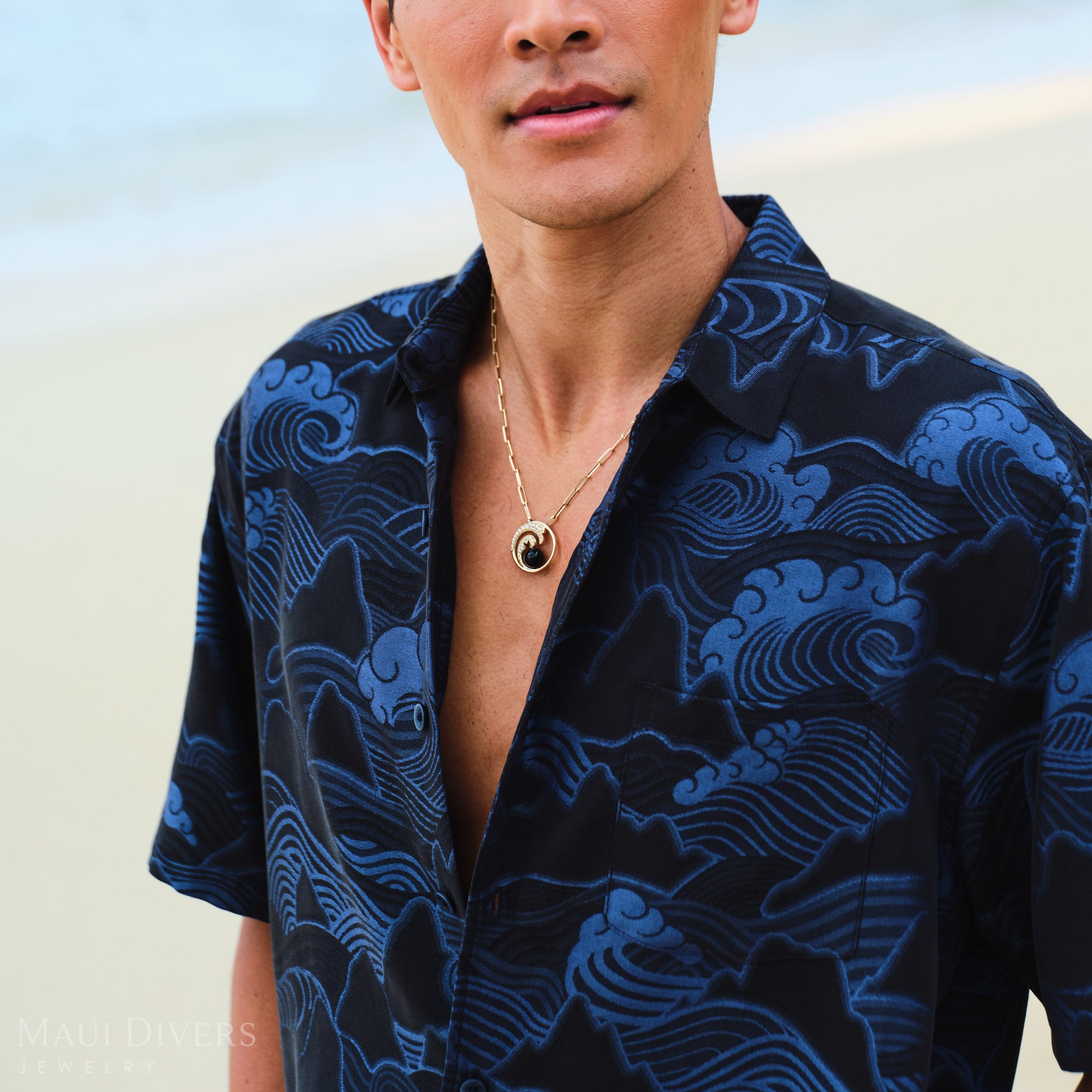 Man wearing a gold necklace with a diamond wave black coral pendant, against a blurred beach background