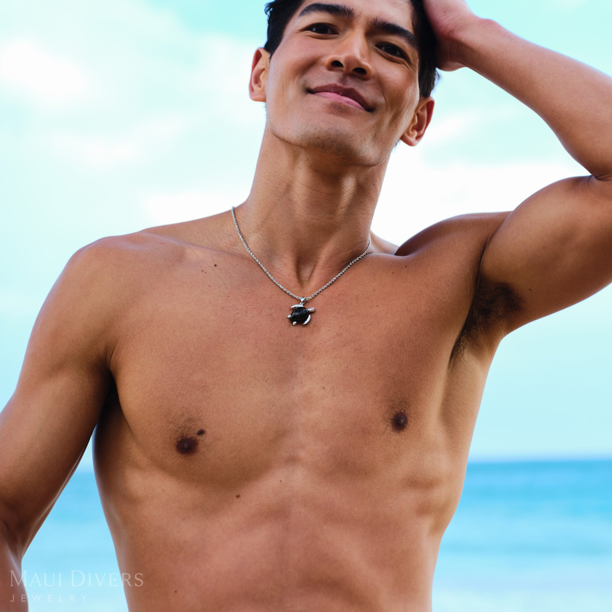 Man wearing a white gold chain with a black coral sea turtle pendant against a blurred ocean background