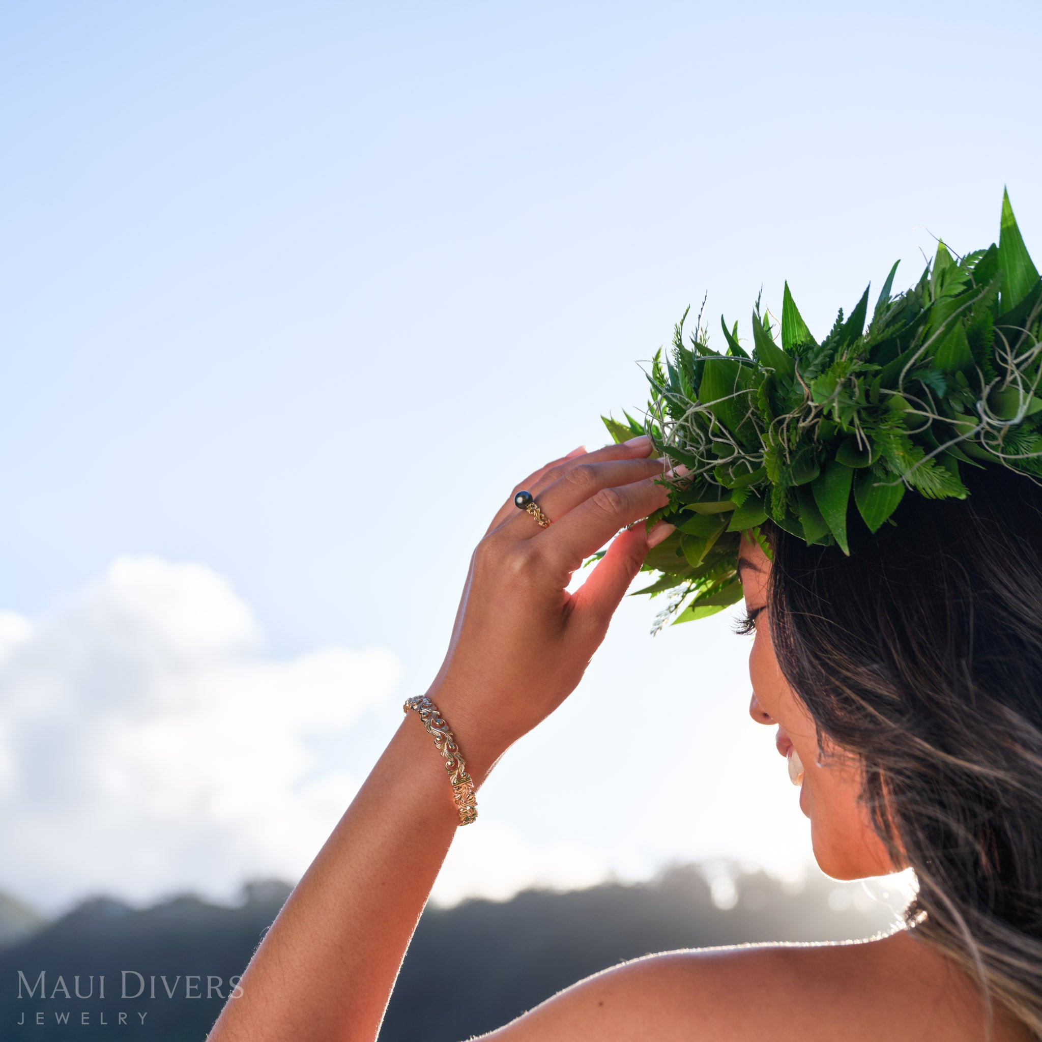 Smiling woman wearing a 14k yellow gold ring with a Tahitian black pearl and 14k yellow gold bracelet against a blurred sky background.